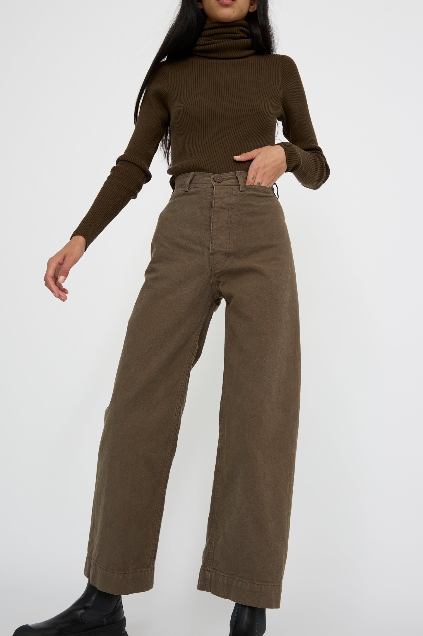 A person wearing Jesse Kamm's Sailor Pant in Mushroom stands against a plain background, pairing them with a brown turtleneck sweater. Their head is not visible.