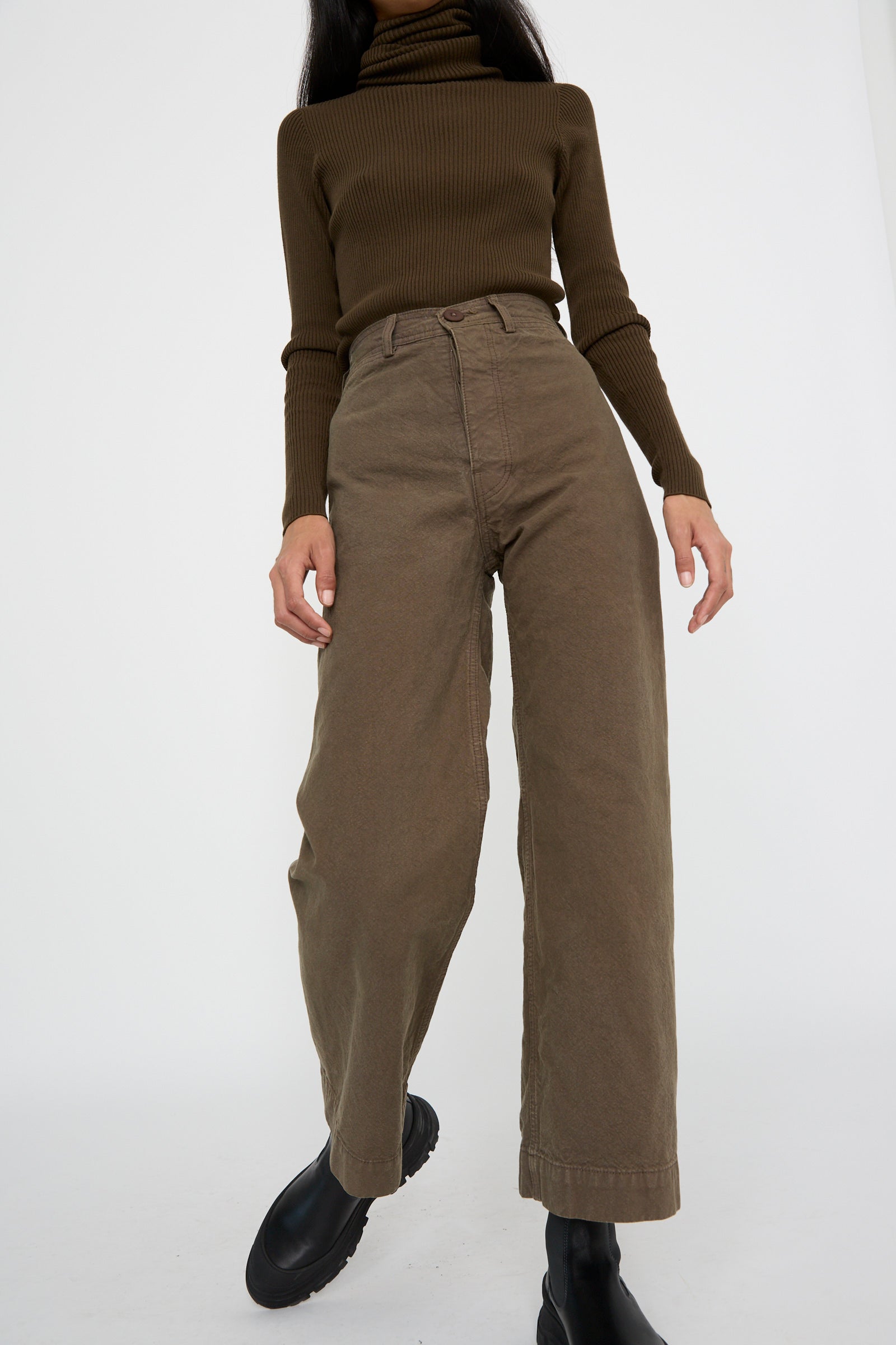 A person wearing the Jesse Kamm Sailor Pant in Mushroom with a brown turtleneck sweater and black boots, posed against a plain white background.