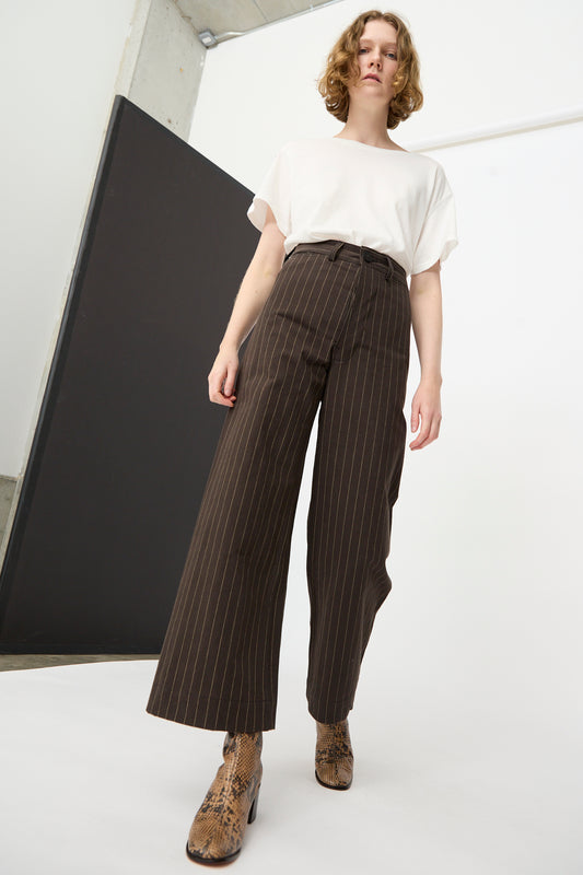 A person wearing a white organic cotton t-shirt, Jesse Kamm’s Sailor Pants in Italian Stripe, and snakeskin boots stands in a minimal studio space.