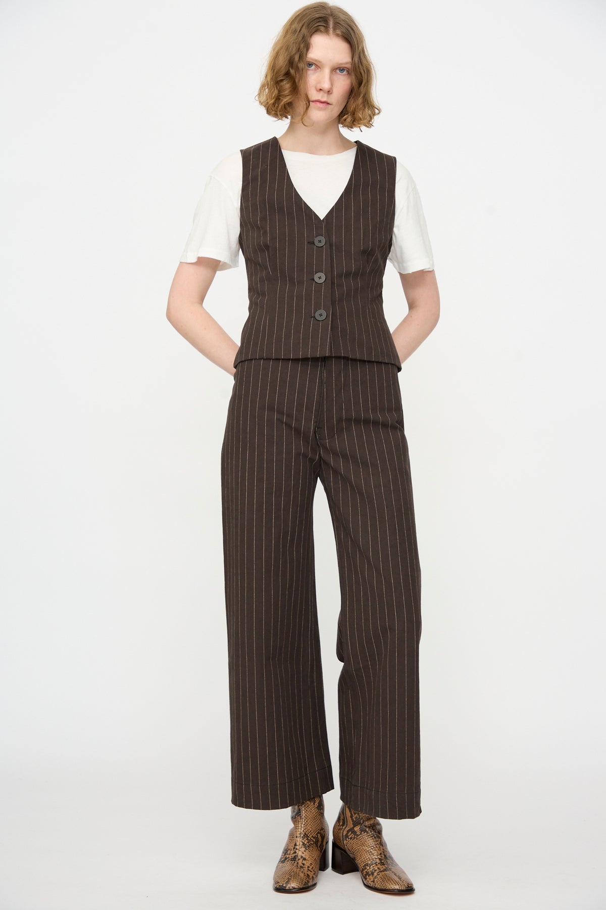 A person wearing the Jesse Kamm Trail Vest in Italian Stripe with matching pants over a white t-shirt, paired with brown snakeskin boots, stands against a plain white background.