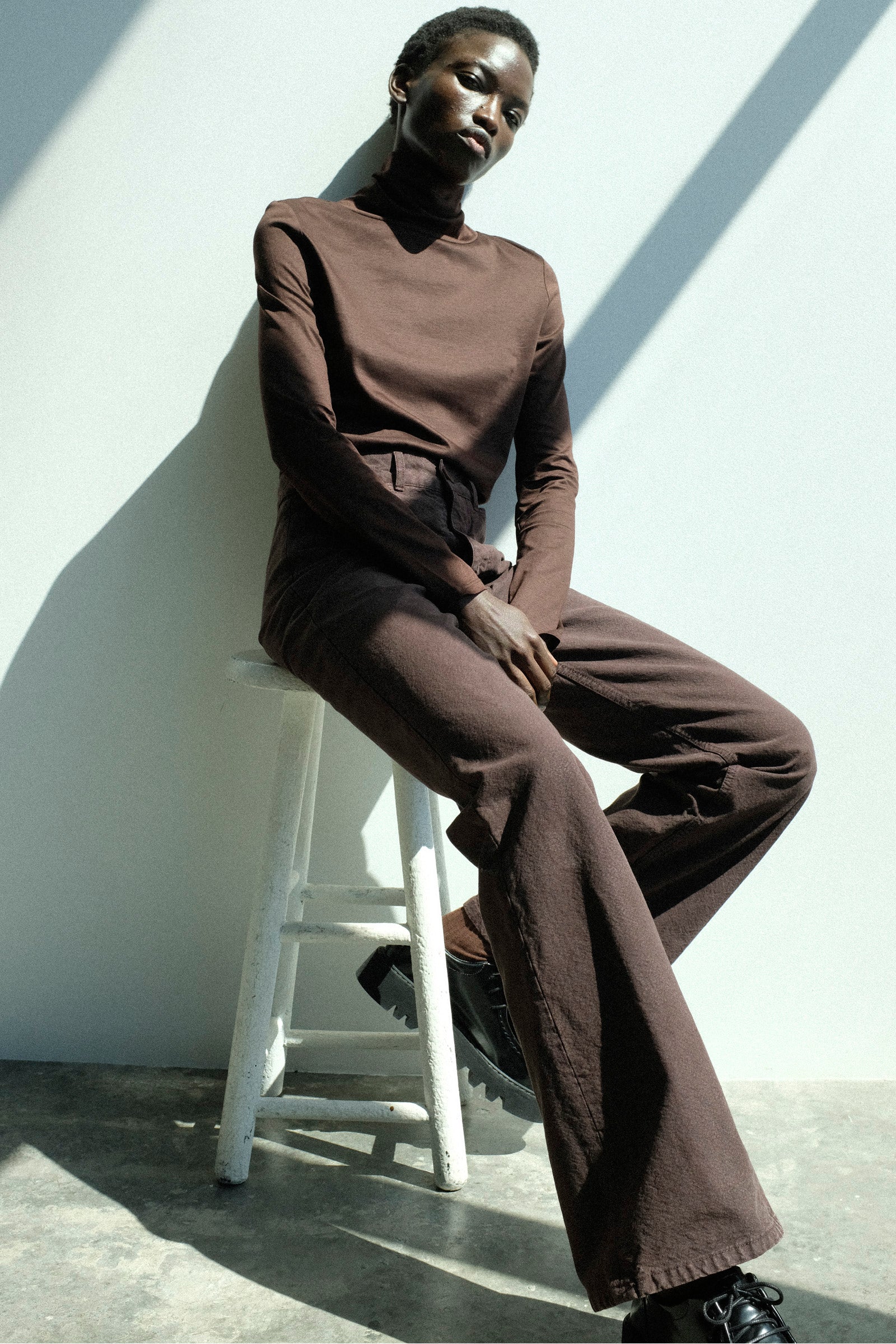Person wearing Jesse Kamm's Japanese Organic Cotton Classic Turtleneck in Raisin with the Organic Canvas Les Deux Pants in Raisin sitting on a white stool against a white wall.