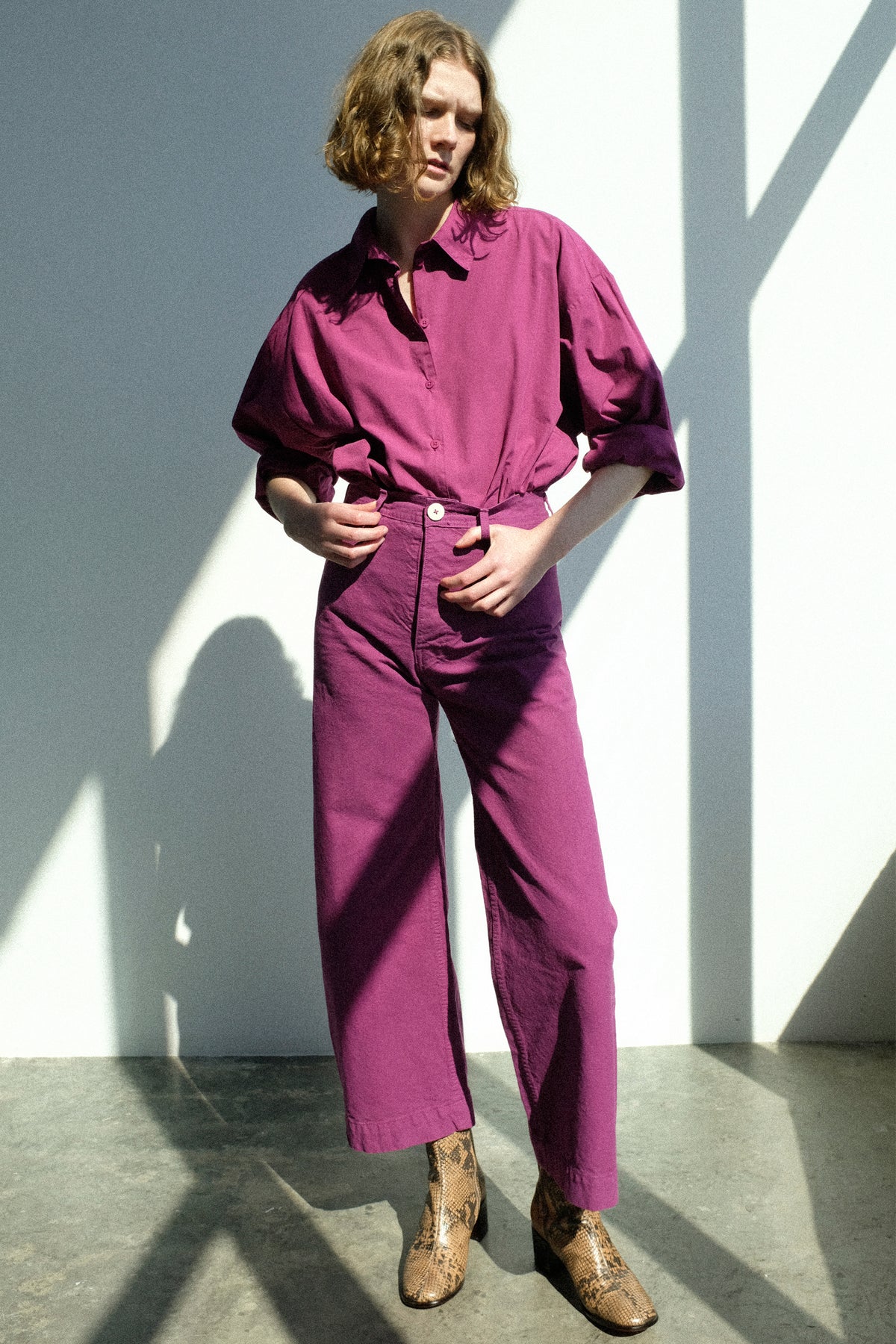 A woman standing against a white wall in sunlight wearing Jesse Kamm's Alberta Shirt in Beet, with the Sailor Pant in Beet.