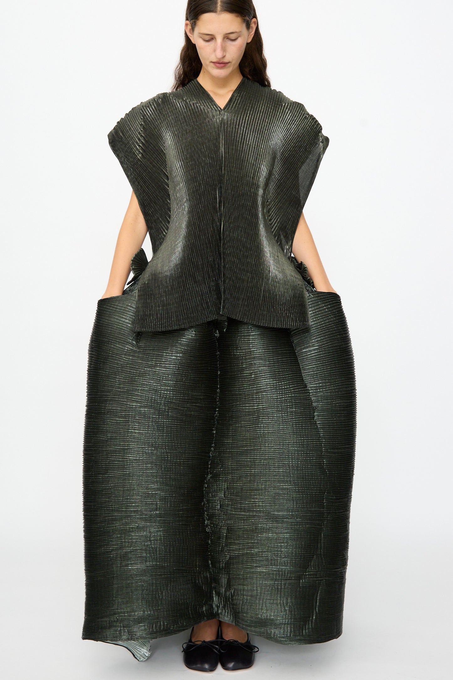 A woman stands against a plain white background, wearing the Julia Heuer Edgar Pleated Poncho in Moss Metallic—a structured, textured dark green piece with wide shoulders and a flared skirt silhouette, her hands in the pockets, evoking the bold proportions of an oversized poncho.