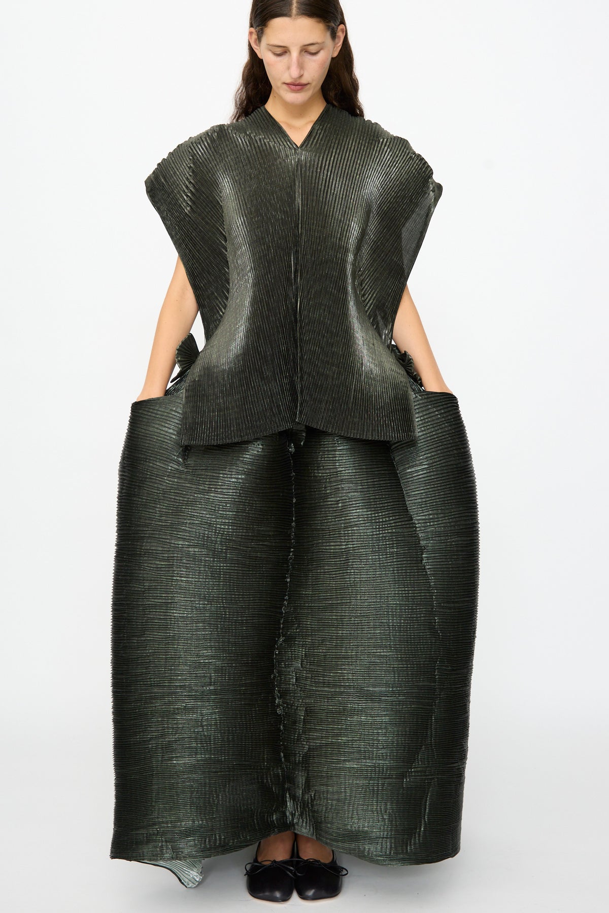 A woman stands against a plain white background, wearing the Julia Heuer Edgar Pleated Poncho in Moss Metallic—a structured, textured dark green piece with wide shoulders and a flared skirt silhouette, her hands in the pockets, evoking the bold proportions of an oversized poncho.