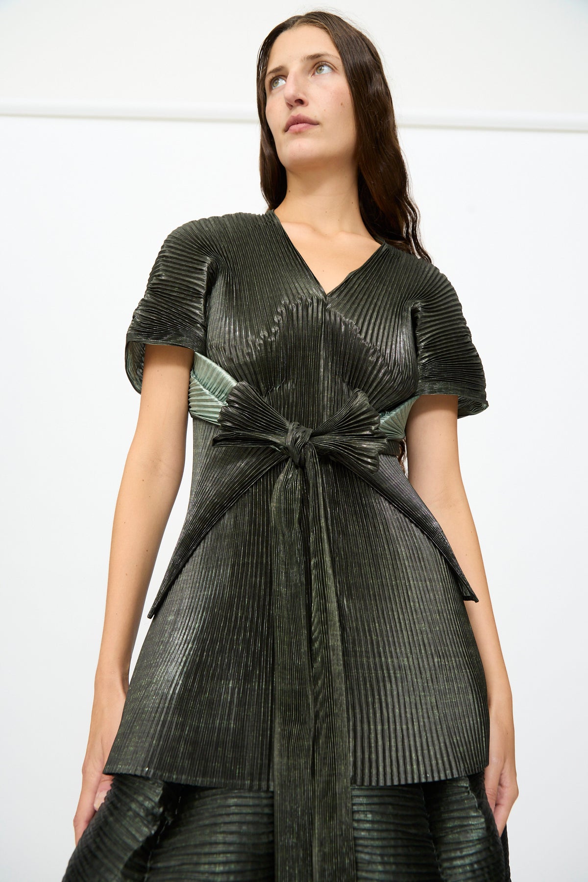 A woman wearing the Julia Heuer Edgar Pleated Poncho in Moss Metallic, featuring a structured silhouette with hand-pleated details and a large waist bow, stands against a plain white background.