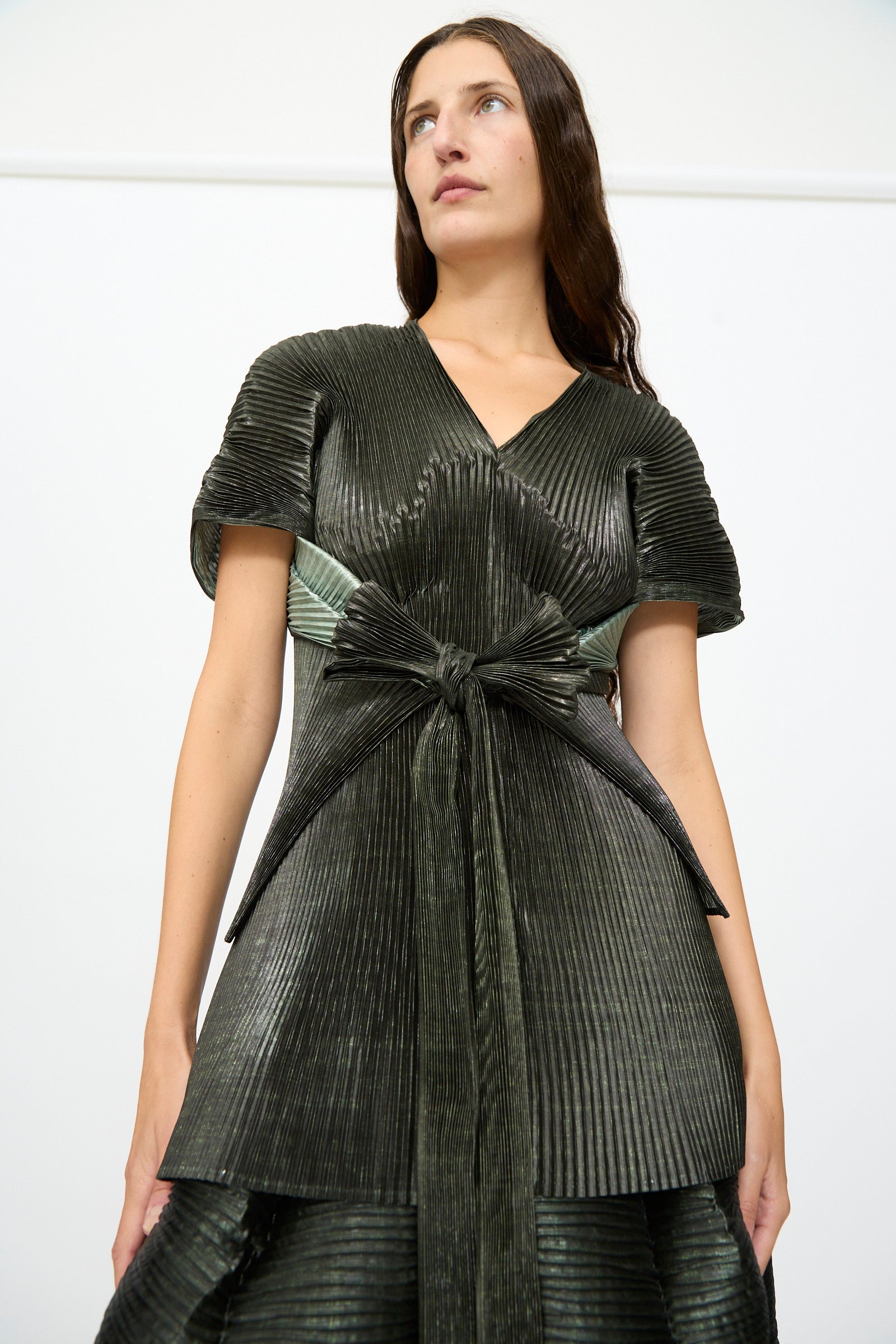 A woman wearing the Julia Heuer Edgar Pleated Poncho in Moss Metallic, featuring a structured silhouette with hand-pleated details and a large waist bow, stands against a plain white background.