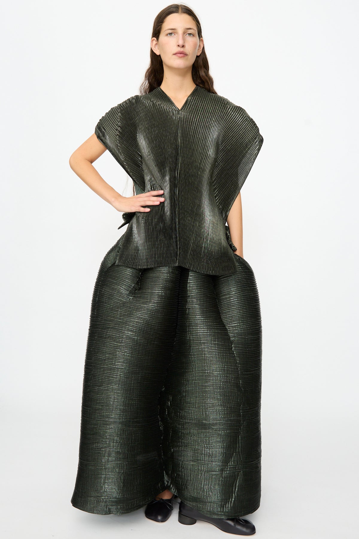 A woman stands against a plain white background, wearing the Julia Heuer Edgar Pleated Poncho in Moss Metallic with a wide matching skirt and black shoes.