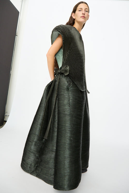 A person stands against a plain background wearing the Julia Heuer Edgar Pleated Poncho in Moss Metallic—a textured, dark green structured top with a hand-pleated finish—paired with a coordinating floor-length skirt.