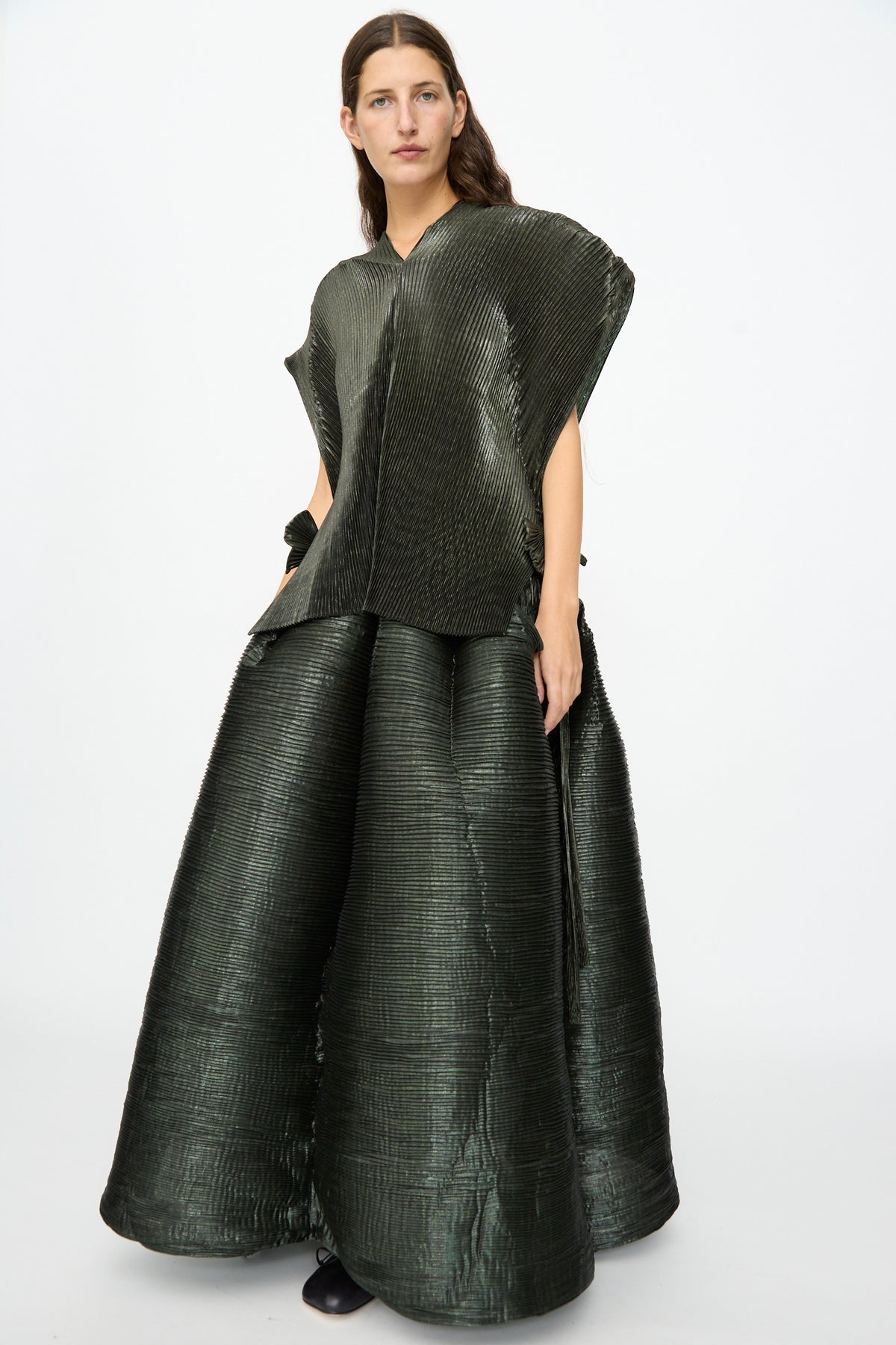 A person wearing the Julia Heuer Edgar Pleated Poncho in Moss Metallic stands against a plain white background, paired with a matching wide, floor-length skirt.