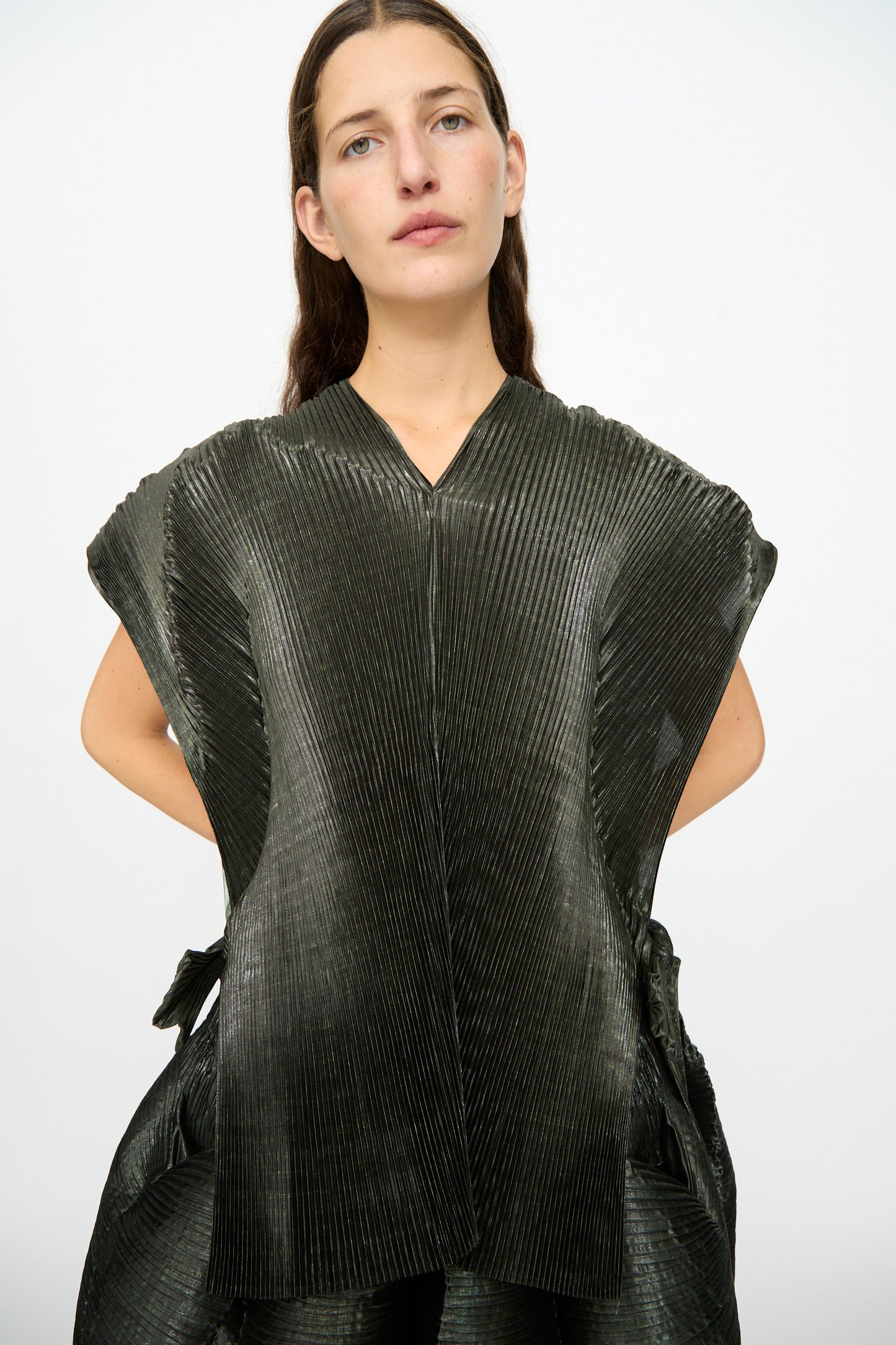 A woman with long brown hair stands against a plain white background, looking directly at the camera, wearing the Edgar Pleated Poncho in Moss Metallic by Julia Heuer—a hand-pleated design featuring wide shoulders.
