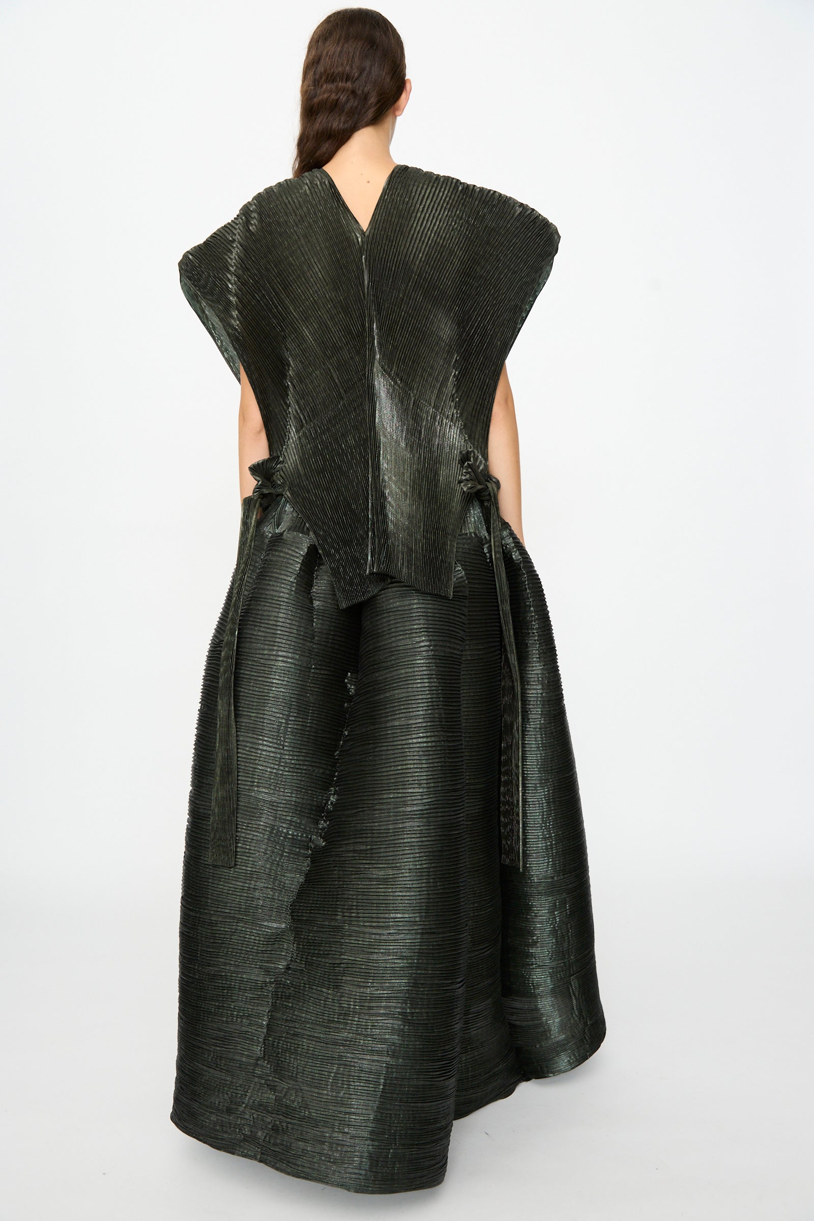 A person is seen from the back against a plain background, elegantly layered in the Julia Heuer Edgar Pleated Poncho in Moss Metallic—an oversized, textured piece with wide shoulders and a long, flowing silhouette. 