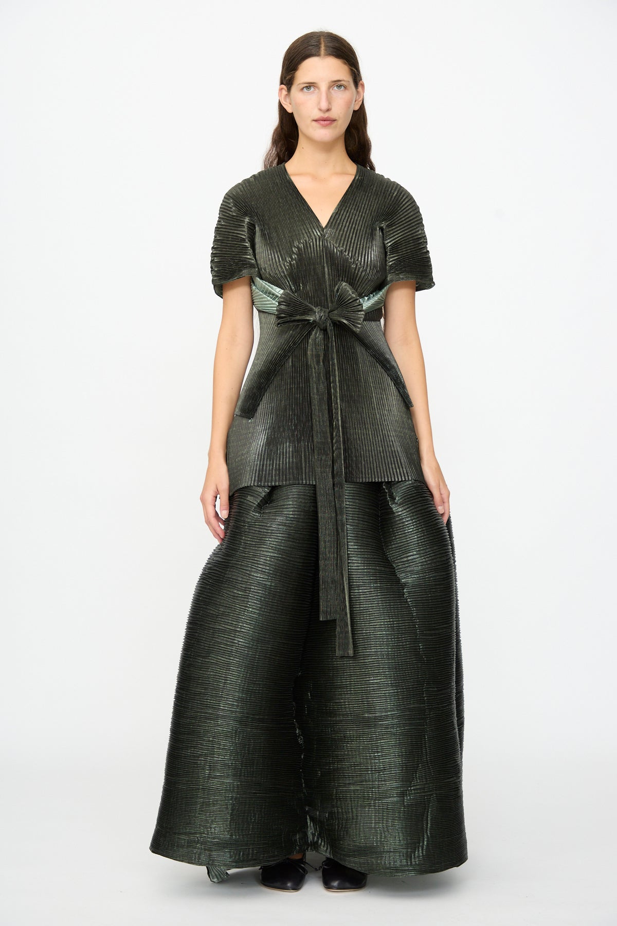 A woman stands against a white background wearing the Edgar Pleated Poncho in Moss Metallic by Julia Heuer, a dark green, hand-pleated metallic piece paired with wide pants and a matching top tied with a large bow at the waist. This striking, structured outfit captures an elegant silhouette and is proudly made in France.