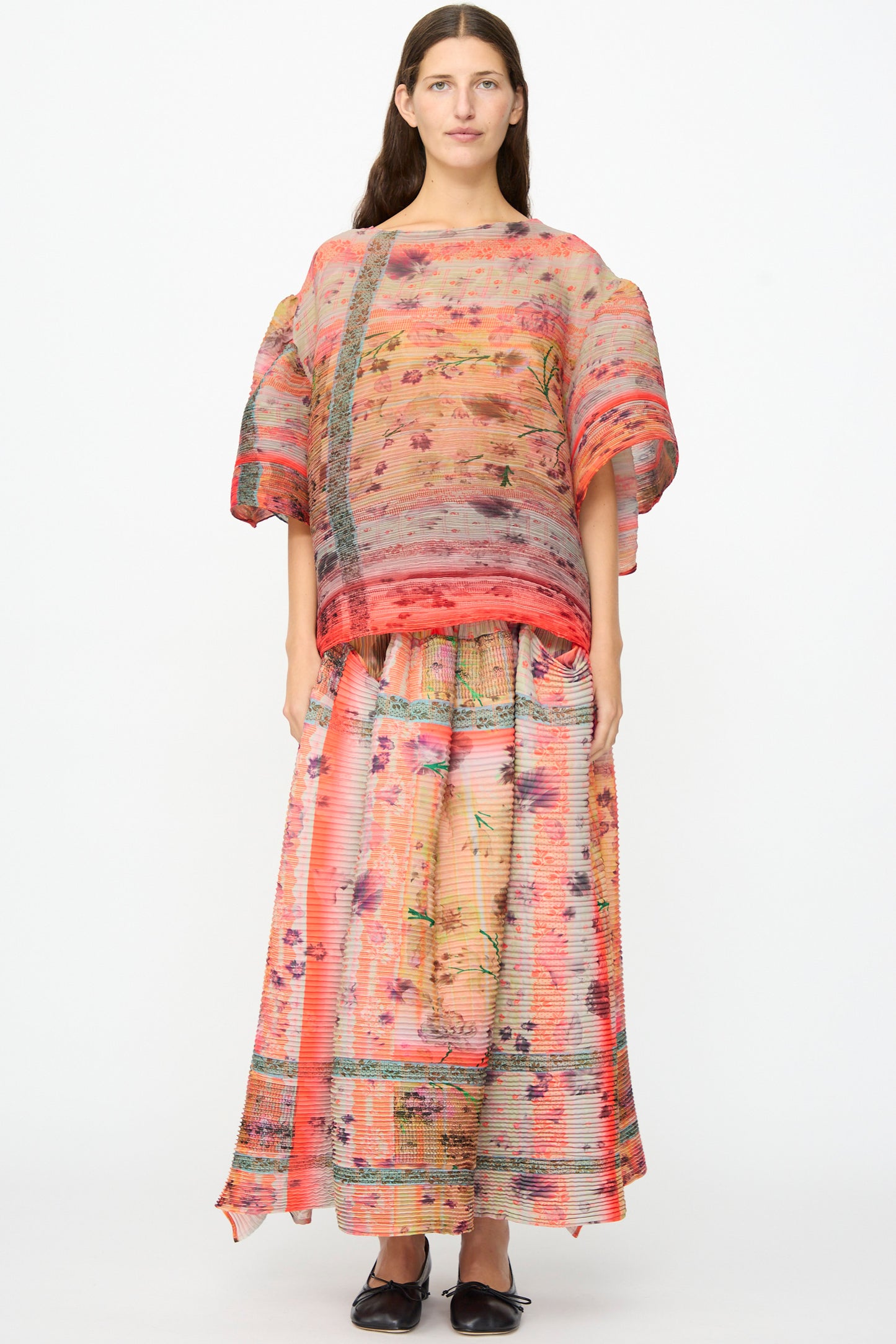 A woman stands against a white background, wearing the Julia Heuer Kenza Handpleated Skirt in Birte—a long, pleated maxi skirt in pink and coral tones crafted from recycled crepe—paired with a loose, sheer floral blouse and black flat shoes.