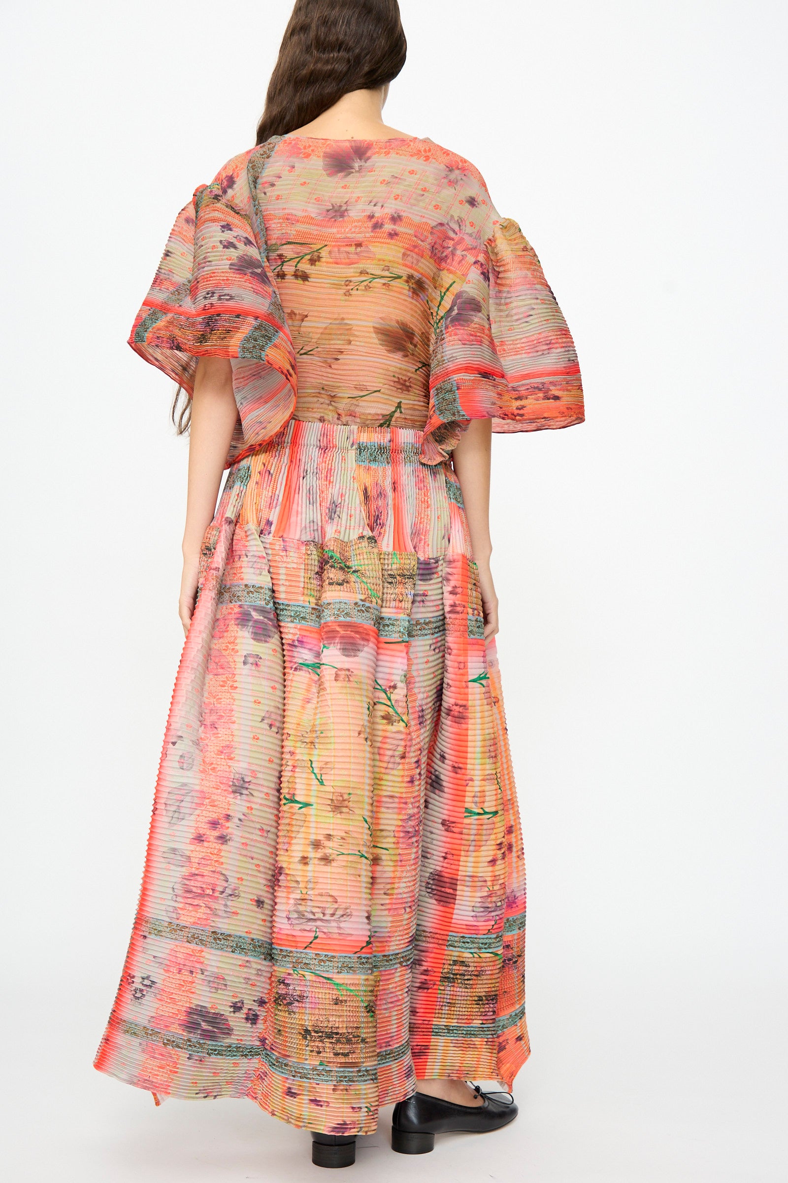 A person stands with their back to the camera, wearing the Julia Heuer Kenza Handpleated Skirt in Birte—a long, pleated, multicolored skirt crafted from recycled crepe—paired with short, wide sleeves and black shoes against a plain background.