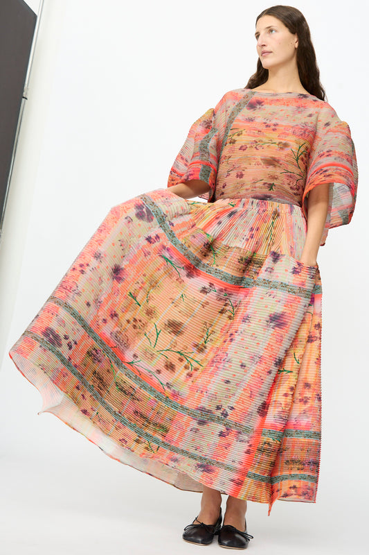 A woman stands against a plain white background and looks to the side, wearing the Julia Heuer Kenza Handpleated Skirt in Birte—a flowing, colorful floral skirt crafted from recycled crepe, styled with wide sleeves.
