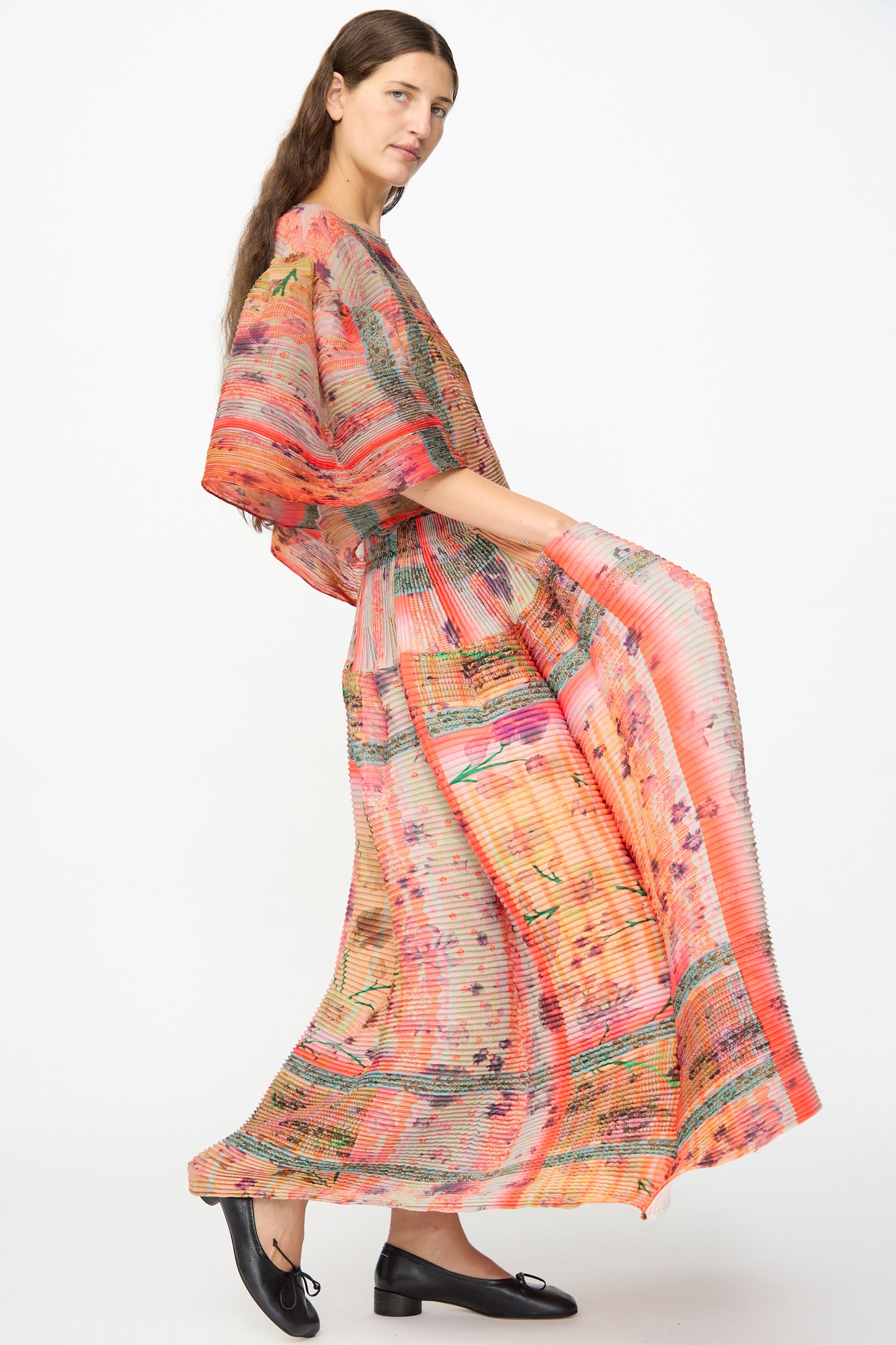 A person stands sideways against a plain white background, wearing the Julia Heuer Kenza Handpleated Skirt in Birte—featuring vibrant floral pleats—and black flats.