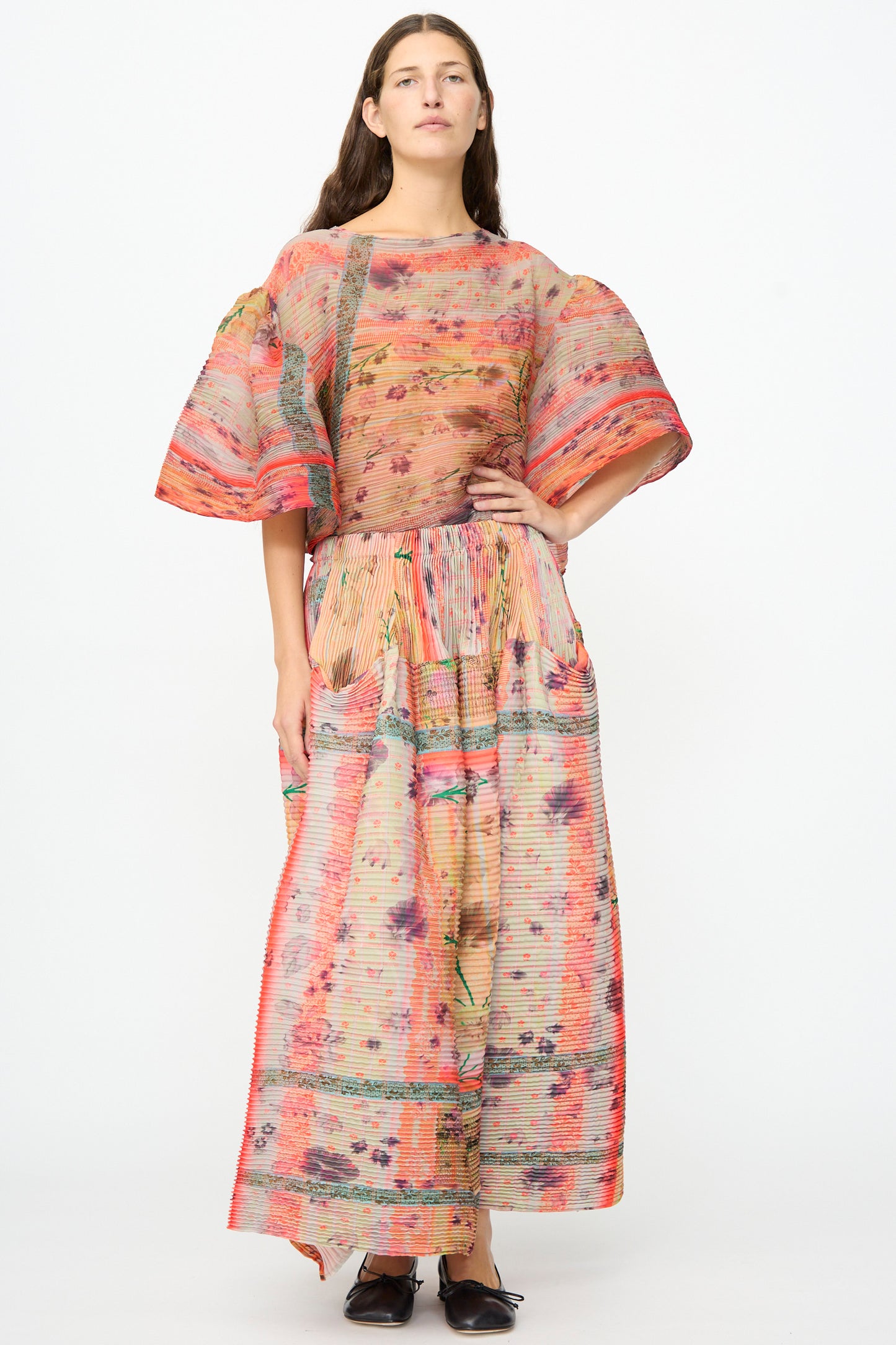 A woman stands wearing the Julia Heuer Kenza Handpleated Skirt in Birte, a vibrant, loose-fitting maxi skirt featuring a colorful floral pattern and crafted from recycled crepe. She pairs the skirt with a coordinating patterned top with wide sleeves and black shoes, while her long brown hair cascades over her shoulders. The background is plain white.