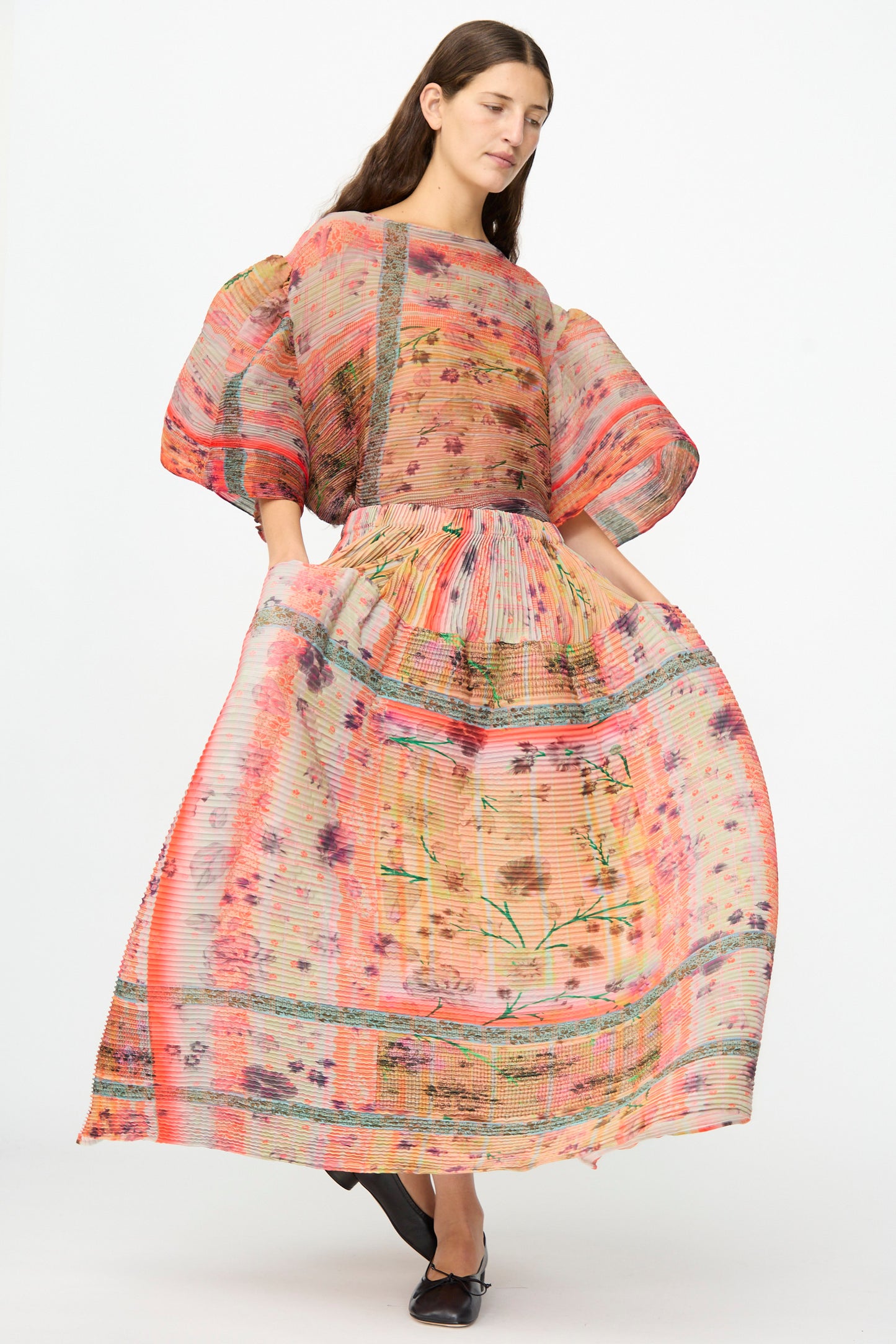 A woman stands against a plain white background wearing the Julia Heuer Kenza Handpleated Skirt in Birte, a long, full-skirted piece crafted from recycled crepe with colorful sheer floral and striped patterns and voluminous sleeves.