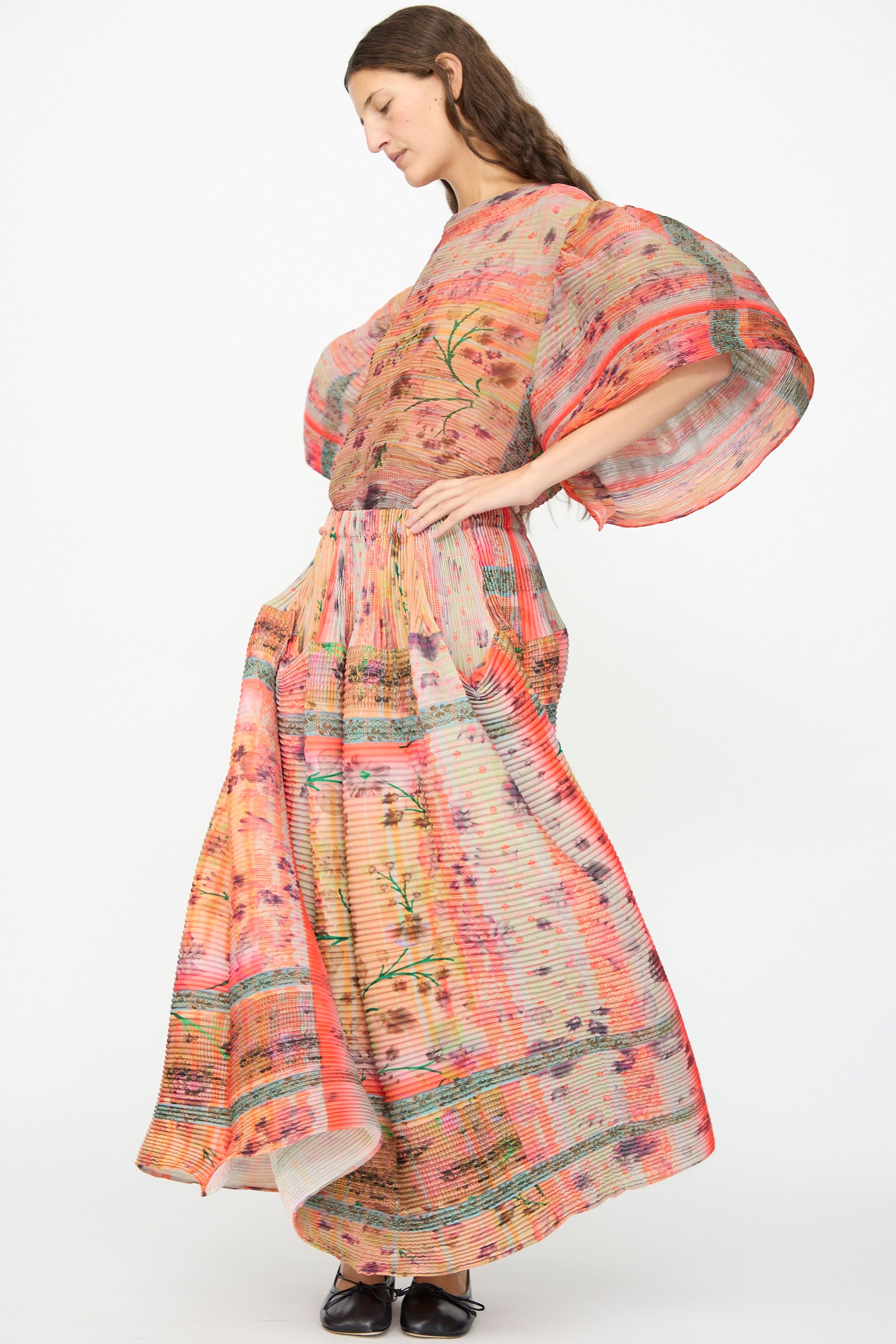 A woman wearing the Julia Heuer Kenza Handpleated Skirt in Birte, which features a flowing, hand-pleated maxi silhouette with a vibrant, multi-colored floral pattern, stands against a plain white background.