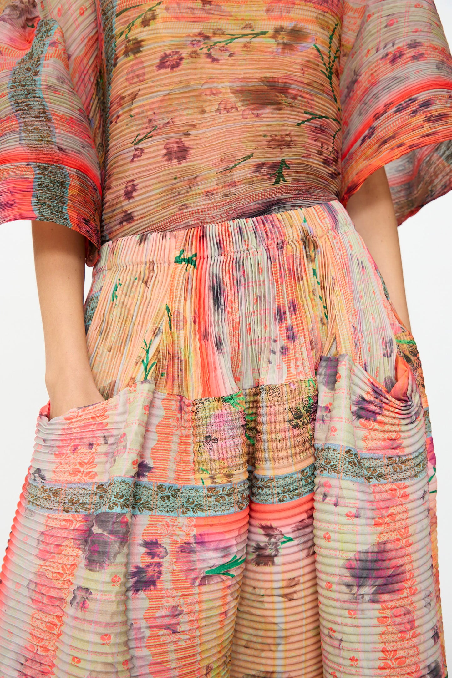 A person wearing the Julia Heuer Kenza Handpleated Skirt in Birte, featuring a colorful floral and stripe pattern, wide sleeves, pleated detailing, hands in pockets, and crafted from recycled crepe.
