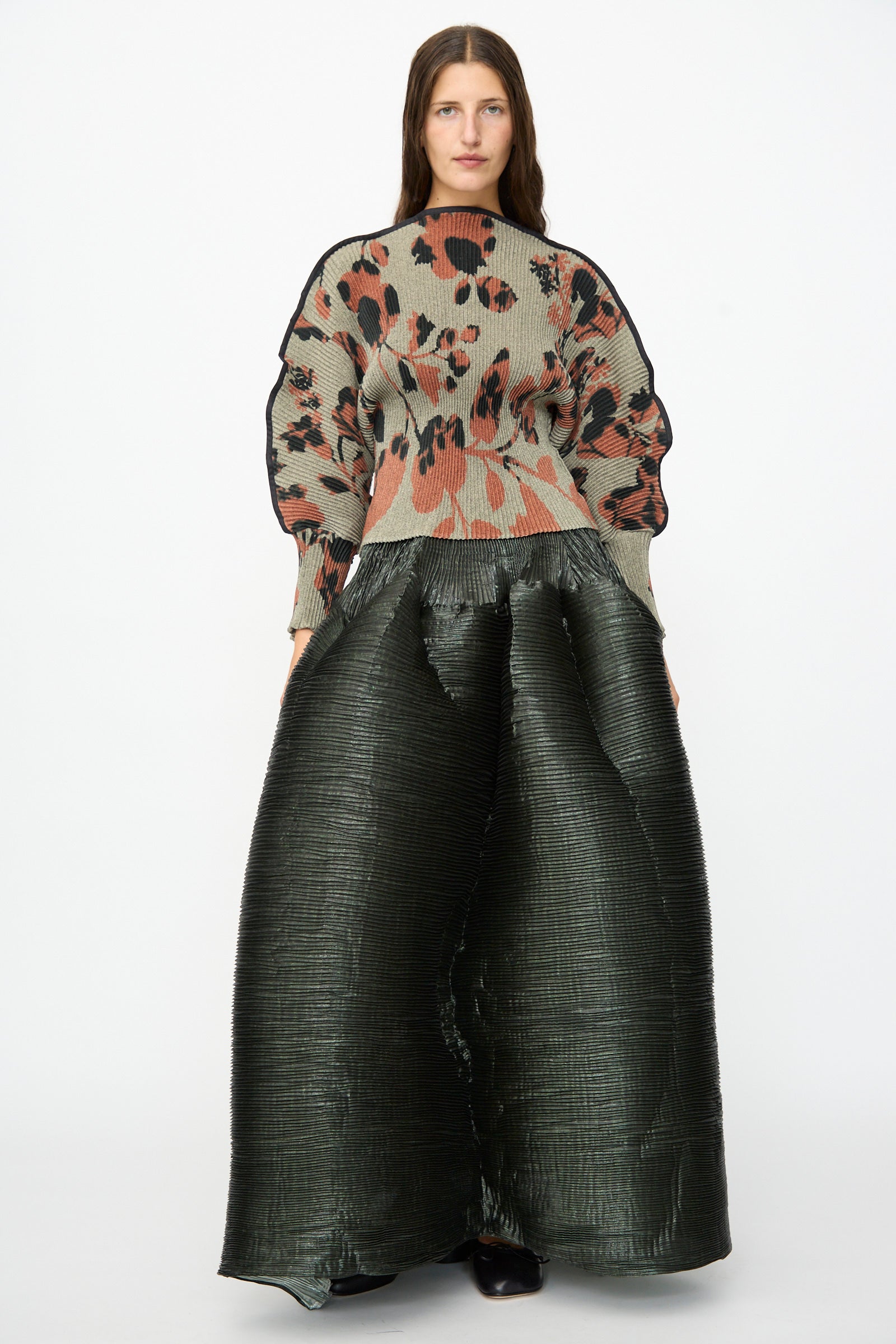 A person stands facing forward against a plain white background, wearing a patterned oversized sweater and the Julia Heuer Kenza Handpleated Skirt in Moss Metallic.