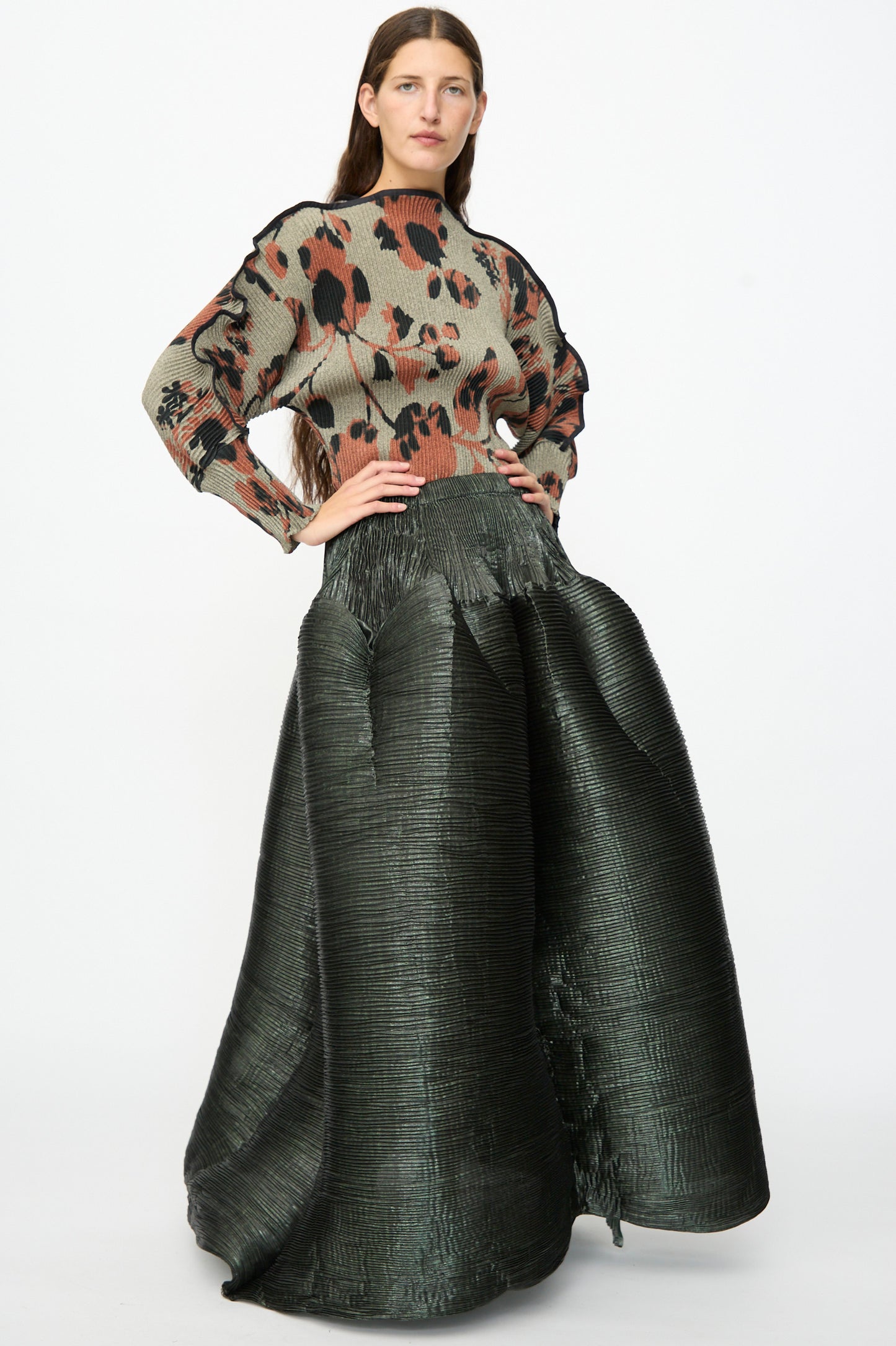 A woman stands against a plain white background wearing the Julia Heuer Kenza Handpleated Skirt in Moss Metallic, paired with a loose, patterned top featuring black and brown abstract shapes.
