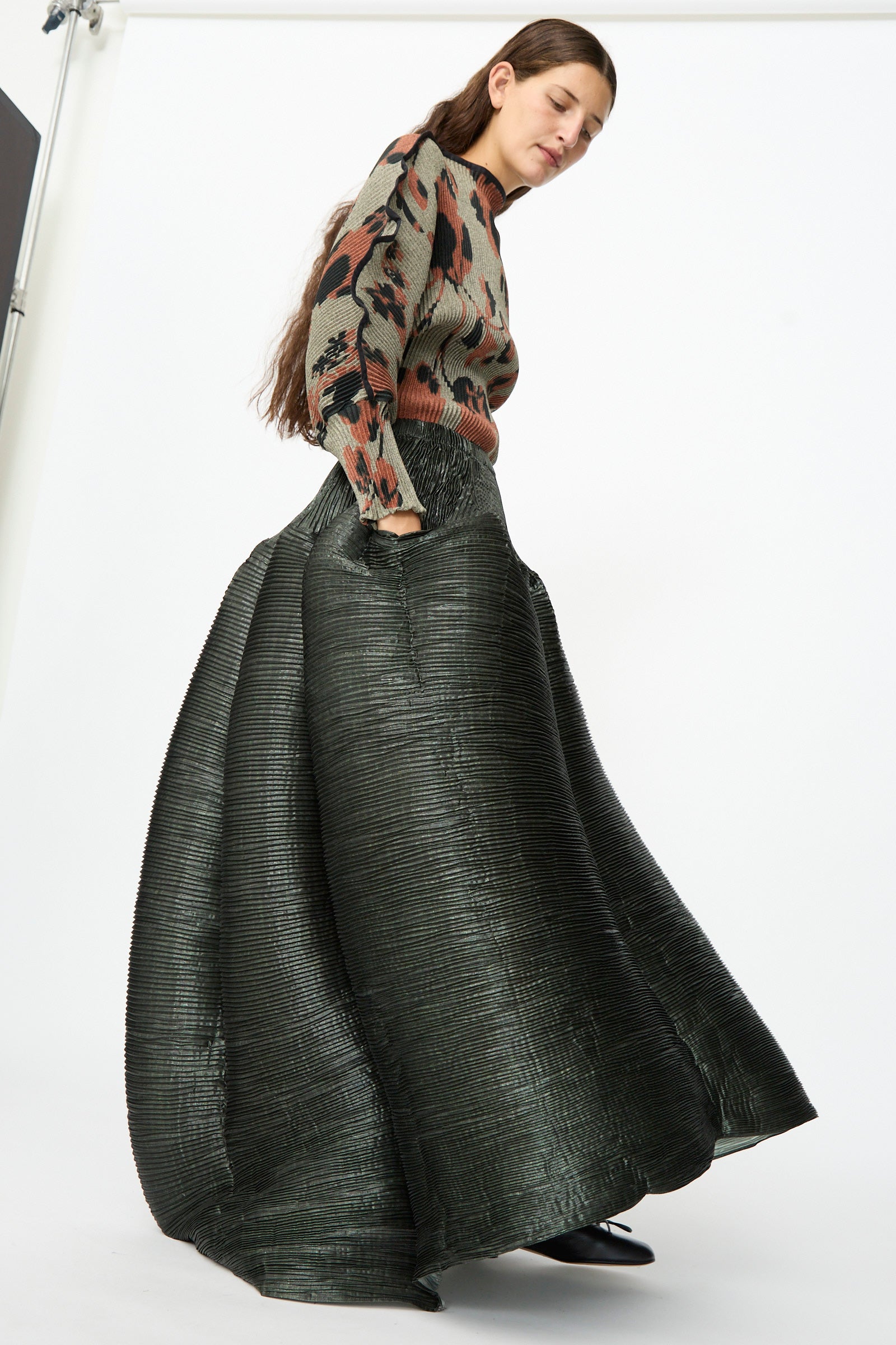 A woman stands against a plain white background, looking downward, wearing a patterned sweater and the Julia Heuer Kenza Handpleated Skirt in Moss Metallic.