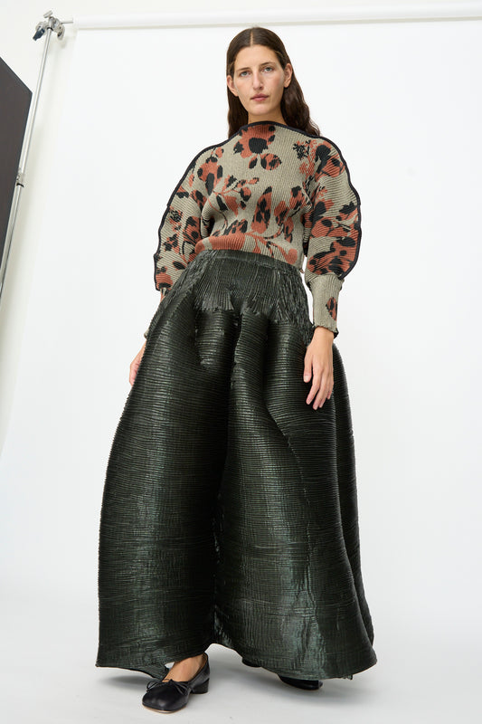 A woman stands against a white backdrop wearing a beige sweater with an abstract dark red and black pattern, paired with the Julia Heuer Kenza Handpleated Skirt in Moss Metallic and black shoes.