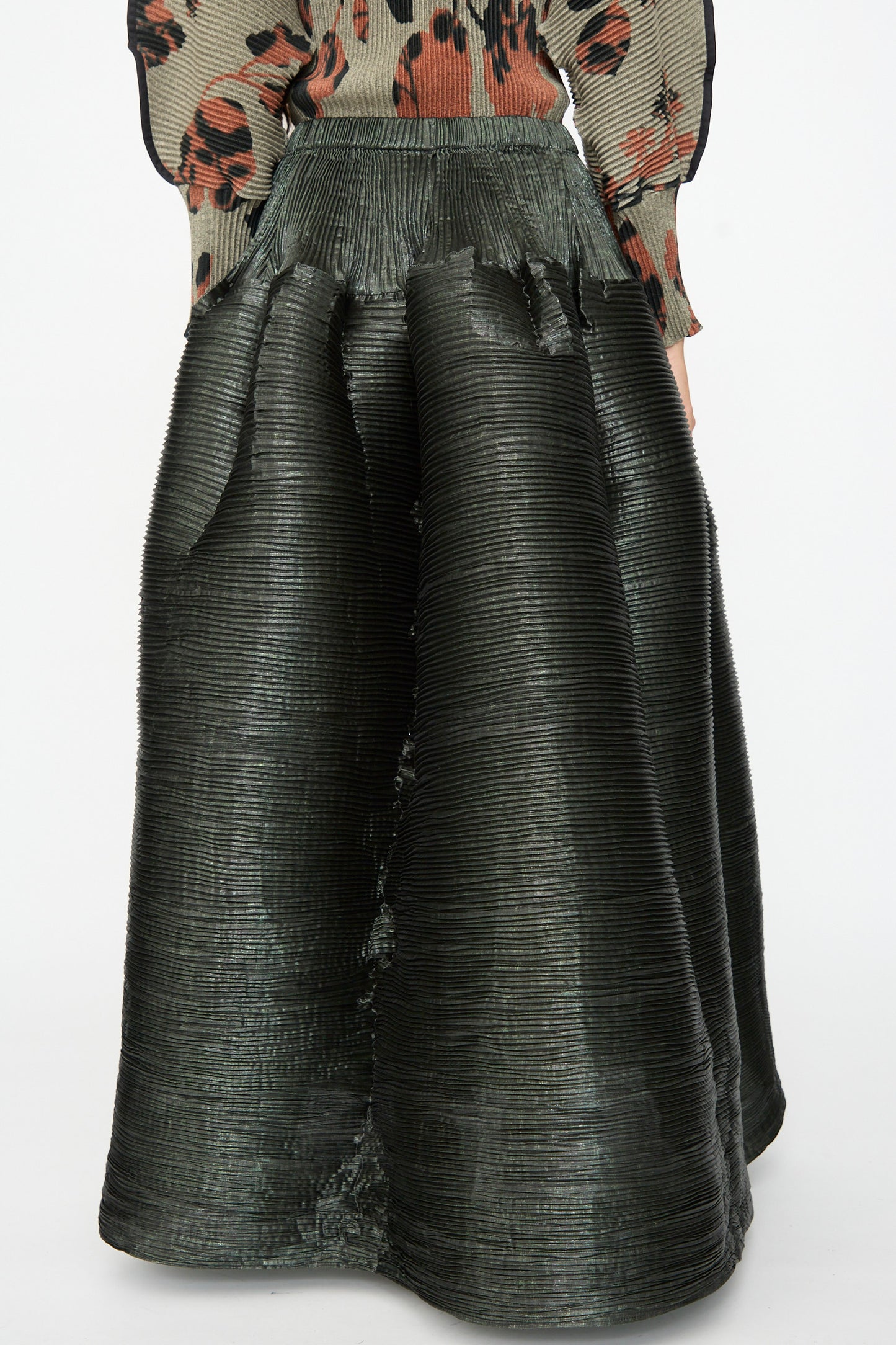 A person wearing the Julia Heuer Kenza Handpleated Skirt in Moss Metallic, featuring elegant high-waisted, hand-pleated detailing, paired with a patterned long-sleeve top.