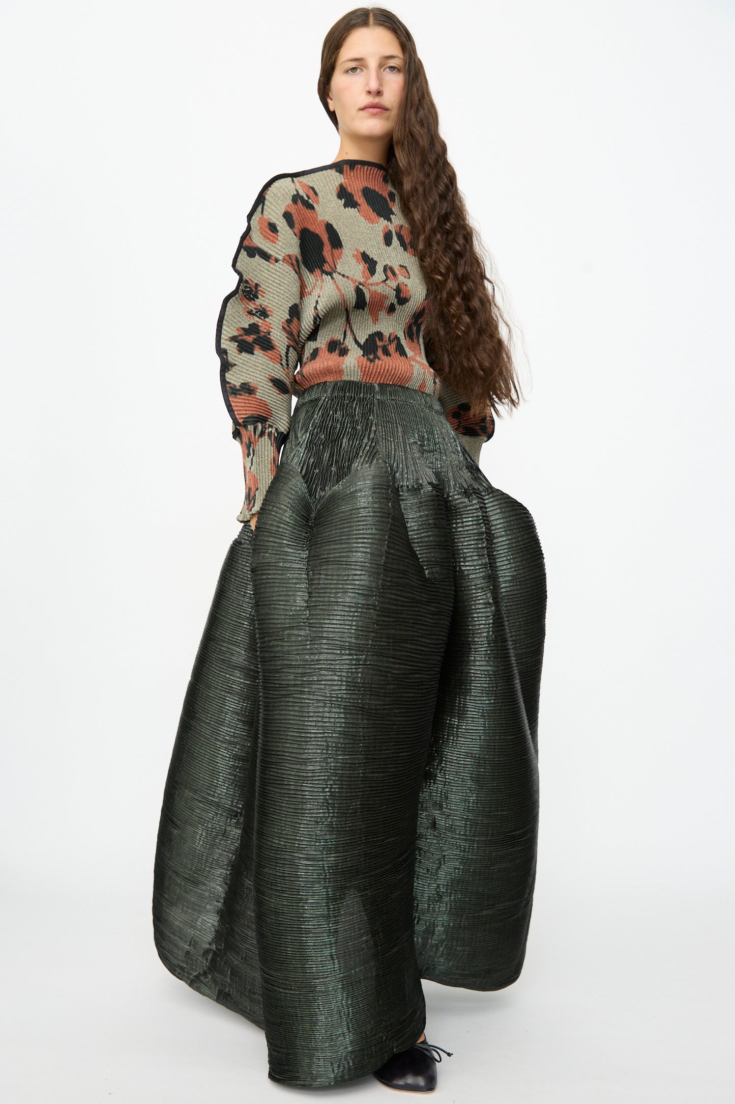 A woman with long brown hair wears a patterned sweater and the Julia Heuer Kenza Handpleated Skirt in Moss Metallic, standing against a plain white background.