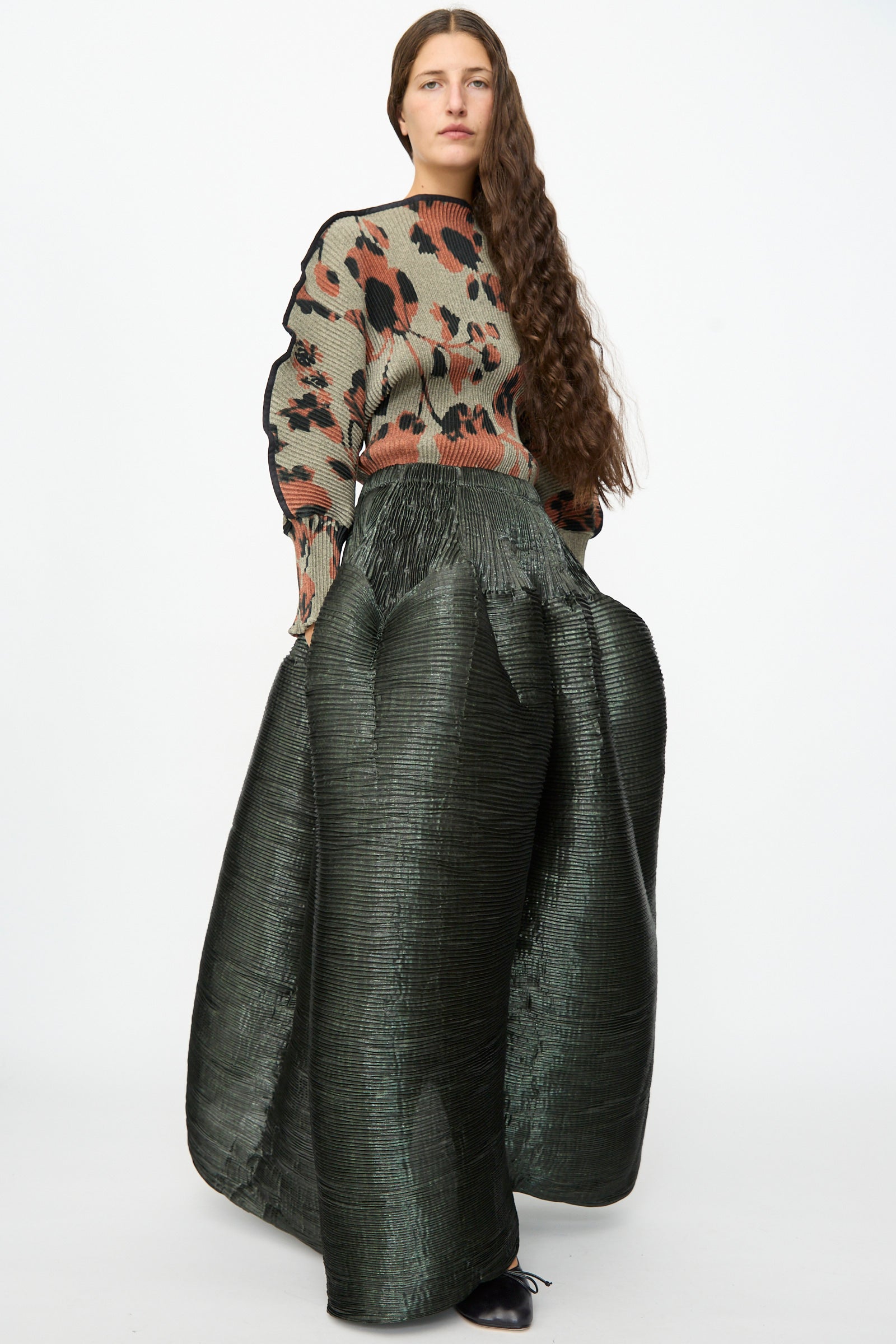 A woman with long brown hair wears a patterned sweater and the Julia Heuer Kenza Handpleated Skirt in Moss Metallic, standing against a plain white background.