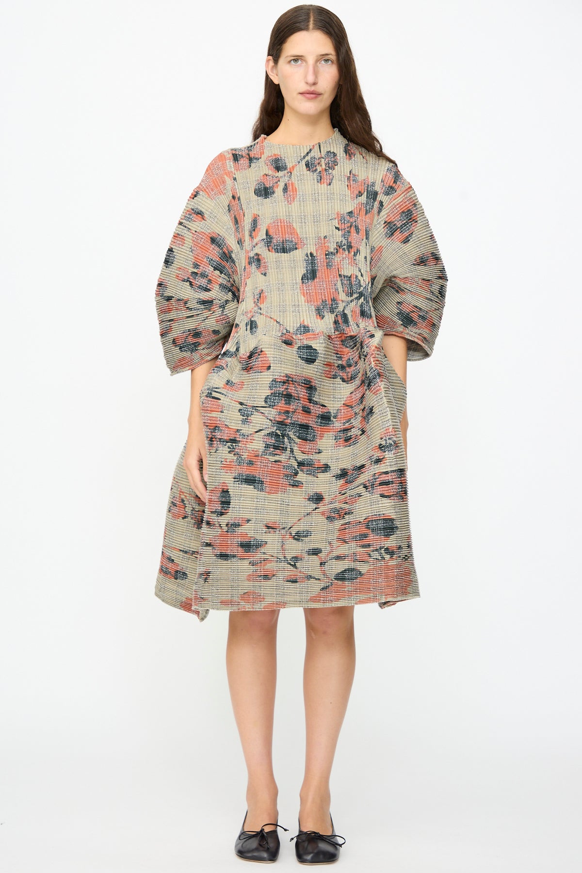 A woman stands facing forward wearing the Julia Heuer Pax Pleated Dress in Sandy Ingrid, which features a sculptural, voluminous silhouette and muted floral pleated fabric. She pairs the look with black pointed-toe flats against a plain white background.