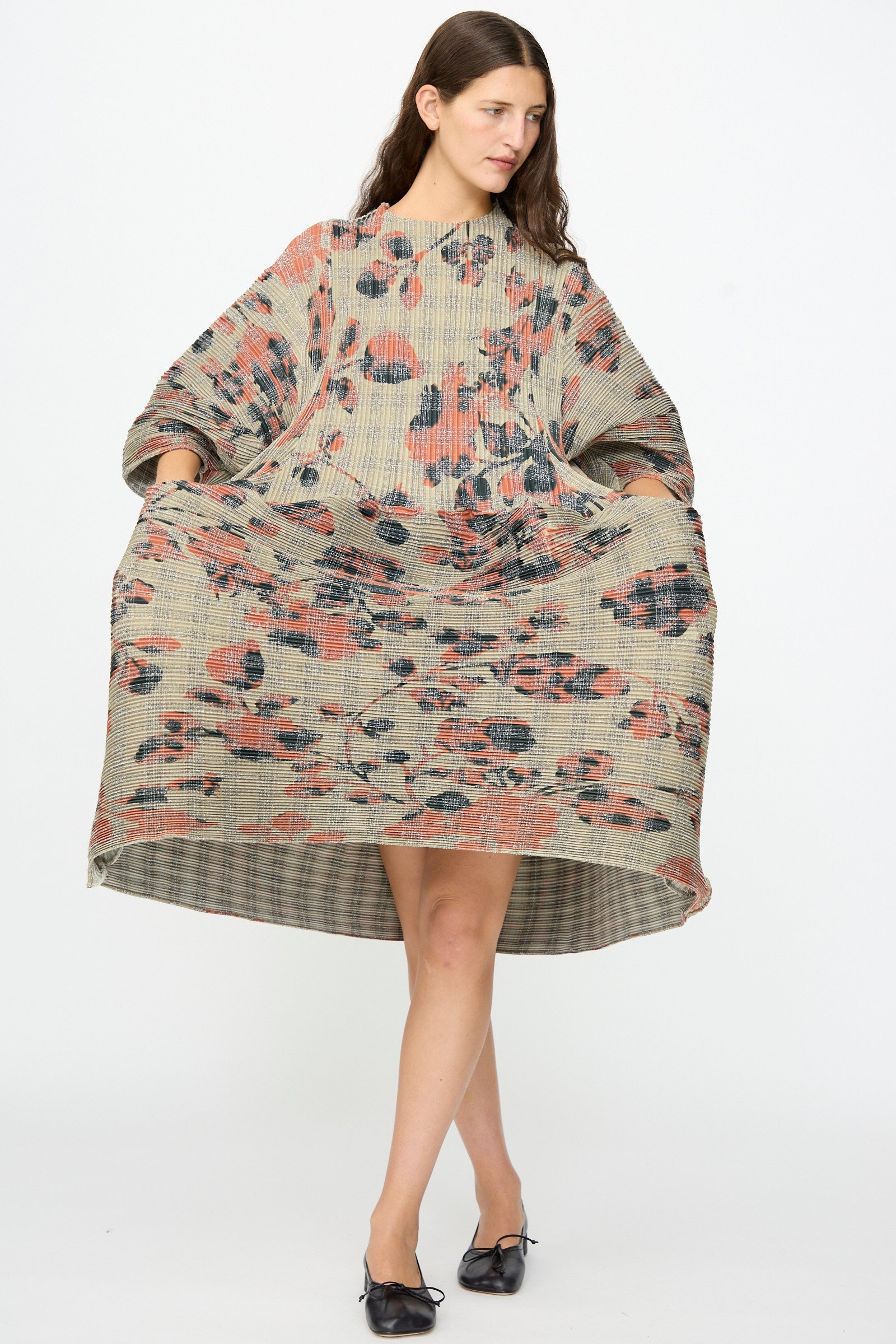 A woman models the Pax Pleated Dress in Sandy Ingrid by Julia Heuer, featuring a loose, textured design in pink, black, and beige. She holds out the sides to highlight its voluminous silhouette while standing against a plain white background.