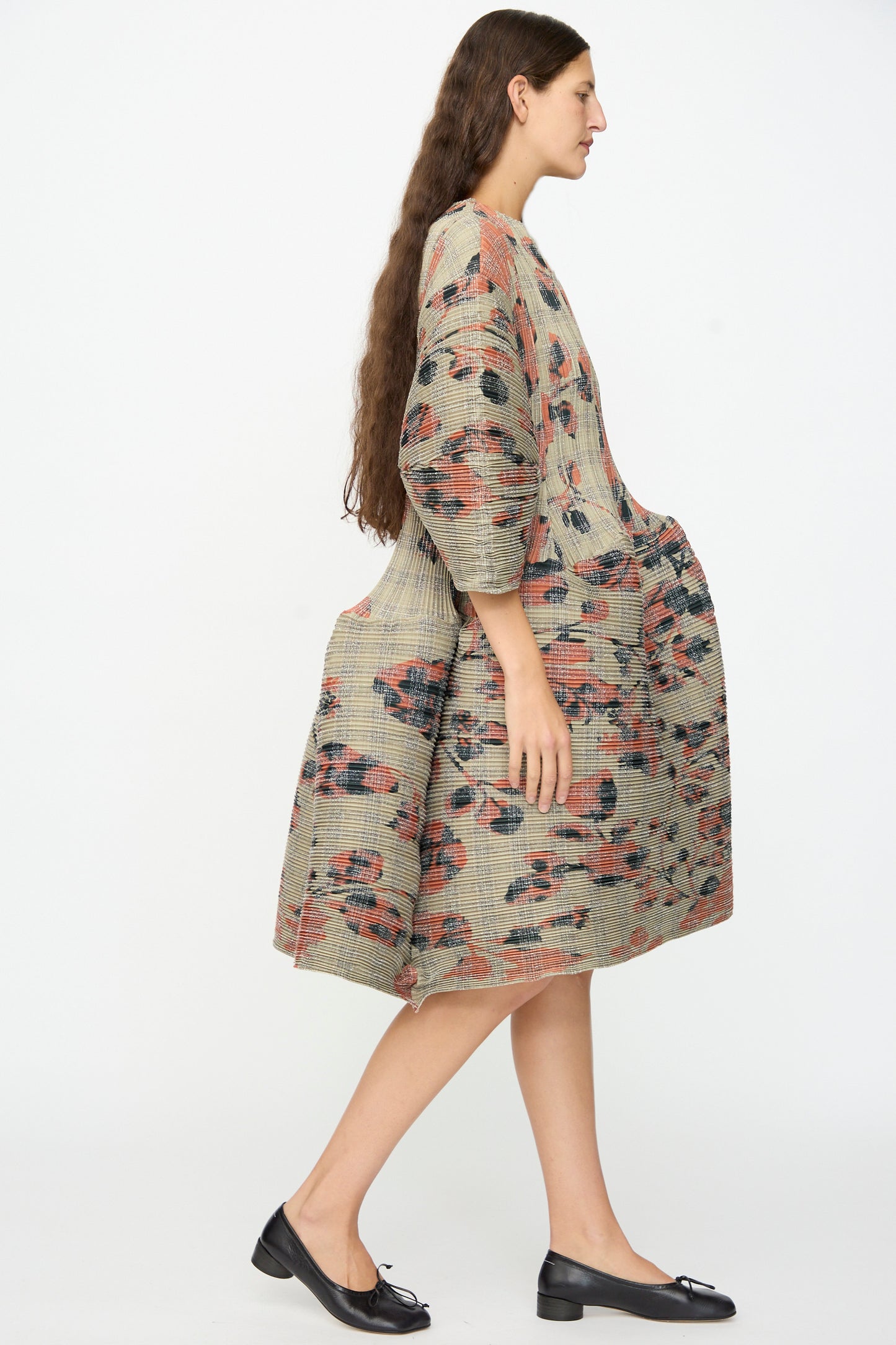A person with long brown hair wears the Julia Heuer Pax Pleated Dress in Sandy Ingrid—a loose, patterned beige dress with three-quarter sleeves—paired with black ballet flats, showcasing a voluminous silhouette as they walk against a plain white background.