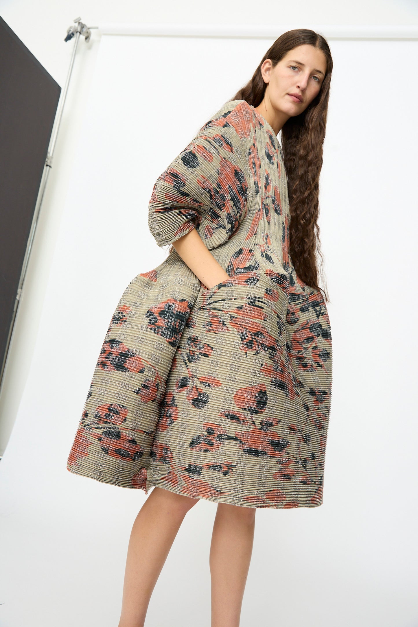 A woman stands in a studio wearing the Julia Heuer Pax Pleated Dress in Sandy Ingrid, featuring a loose, voluminous silhouette and a textured floral pattern. She has long brown hair and her hands are tucked into the dress pockets.