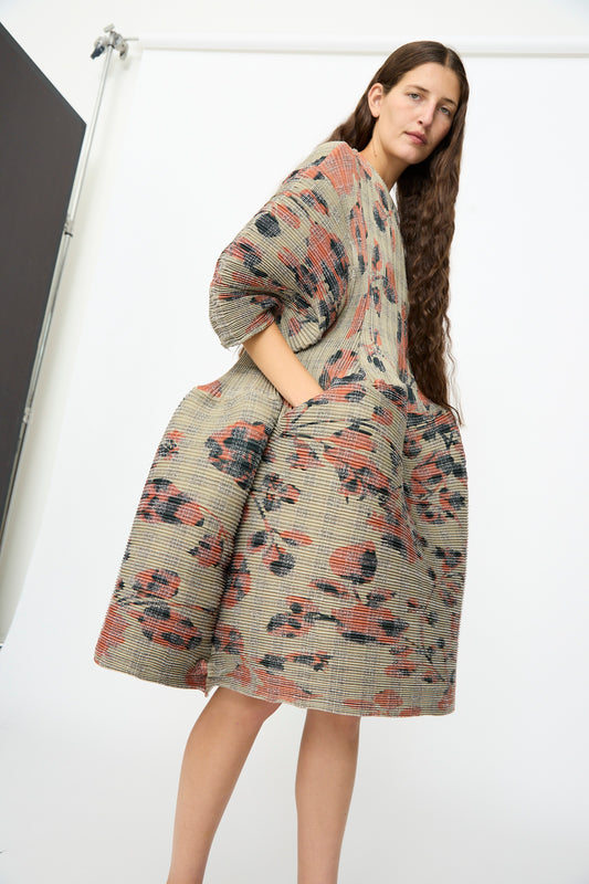 A woman stands in a studio wearing the Julia Heuer Pax Pleated Dress in Sandy Ingrid, featuring a loose, voluminous silhouette and a textured floral pattern. She has long brown hair and her hands are tucked into the dress pockets.