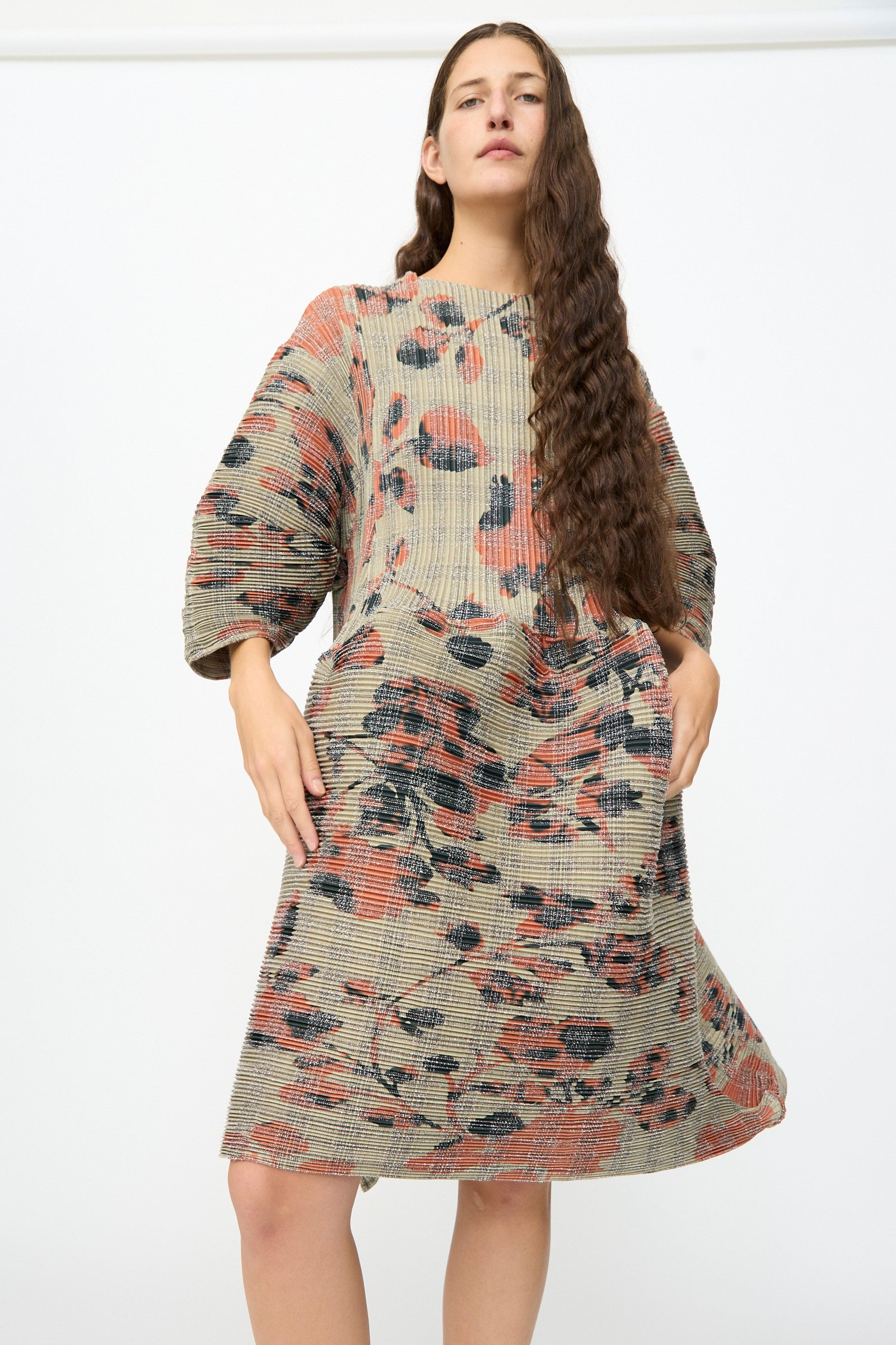 A person wearing the Julia Heuer Pax Pleated Dress in Sandy Ingrid—a loose-fit, knee-length dress with a muted plaid and floral pattern, featuring a voluminous silhouette reminiscent of a sculptural midi dress—stands against a plain white background.