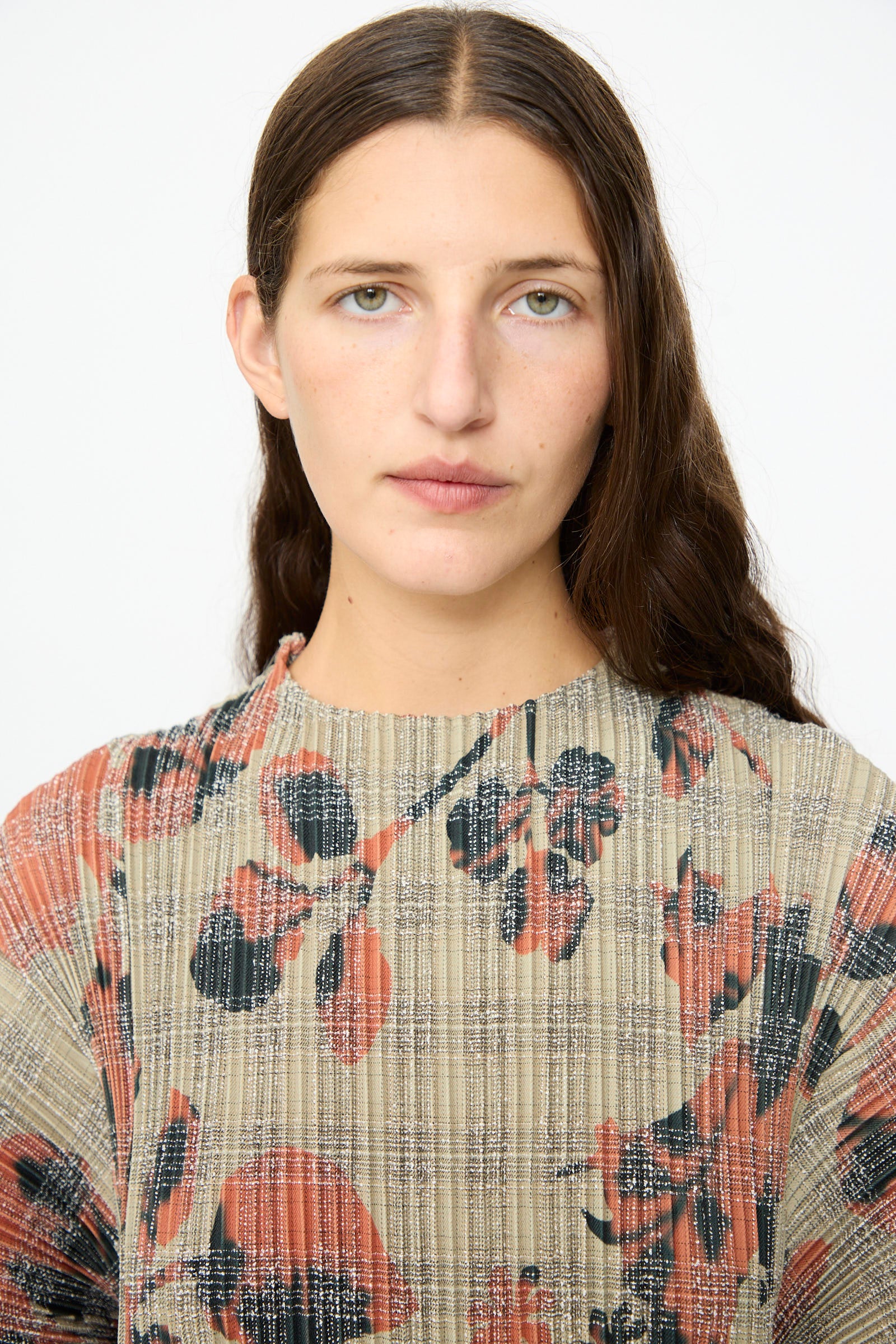 A person with long brown hair and green eyes wears the Julia Heuer Pax Pleated Dress in Sandy Ingrid, featuring pleated fabric with red and black floral and plaid patterns, standing against a plain white background.