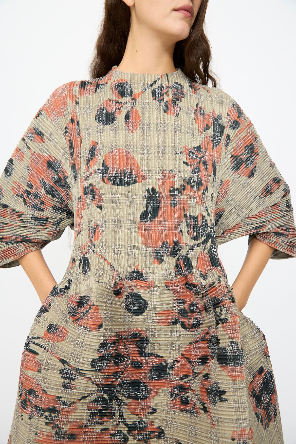A woman wears the Julia Heuer Pax Pleated Dress in Sandy Ingrid, featuring a voluminous silhouette and textured pleats with a plaid base adorned by an abstract red and black floral pattern, standing against a plain background.
