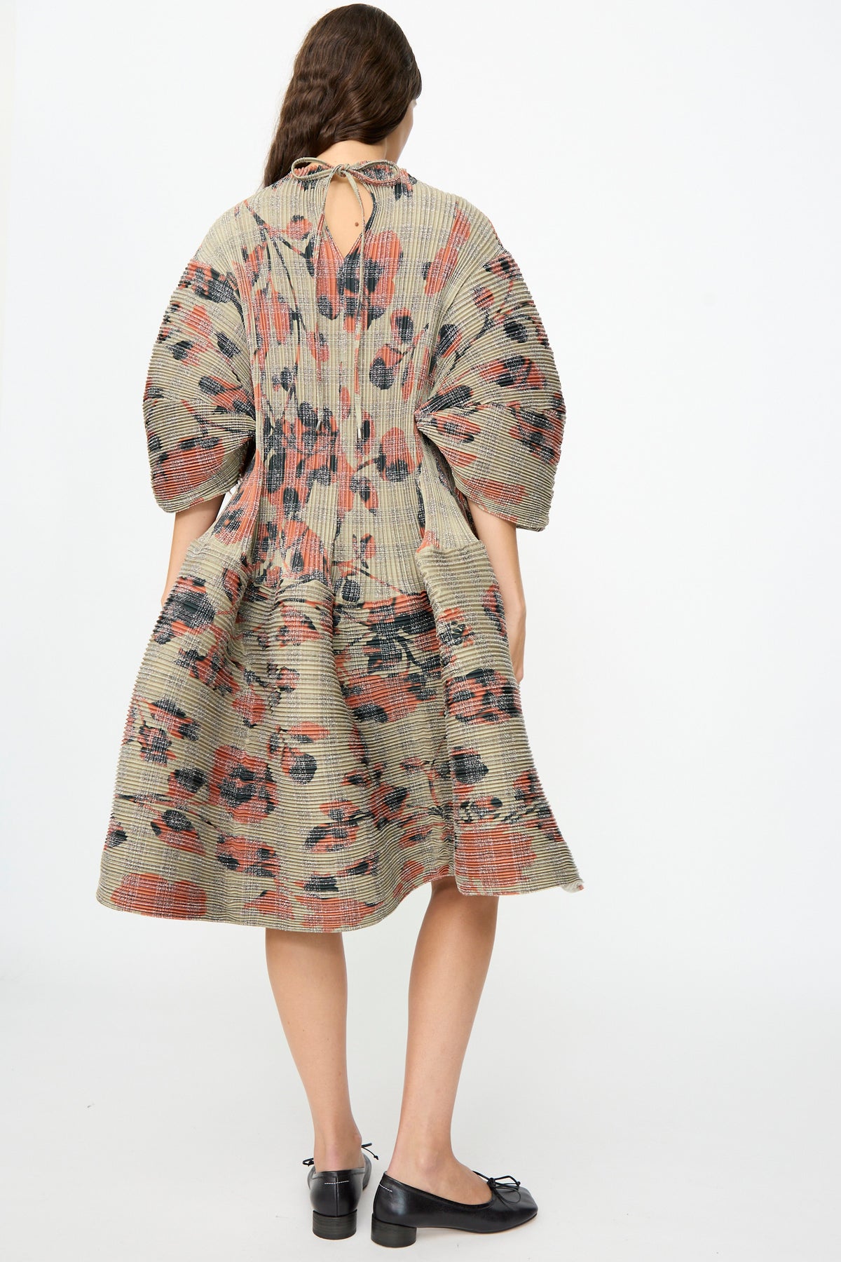 A woman stands facing away, dressed in the Julia Heuer Pax Pleated Dress in Sandy Ingrid—a sculptural midi design featuring a loose, voluminous silhouette crafted from pleated floral fabric with billowy sleeves— paired with black flat shoes.