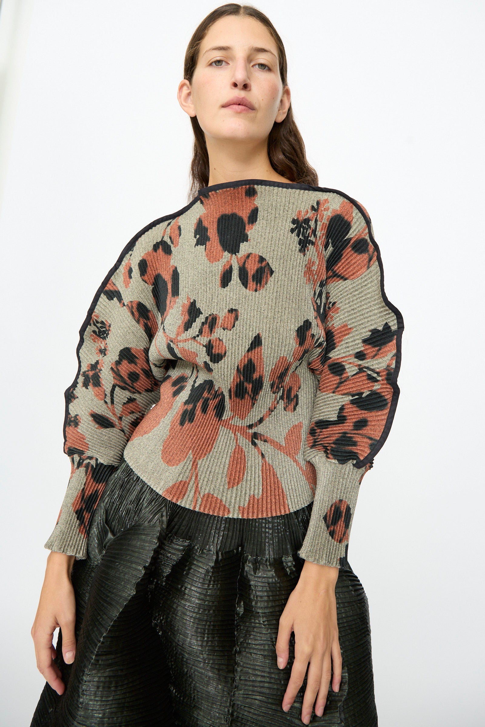 A woman wears the Julia Heuer Radok Pleated Knit Pullover in Ingrid—featuring abstract floral patterns in earthy tones and ribbed long sleeves—with a black textured skirt, standing against a plain white background.