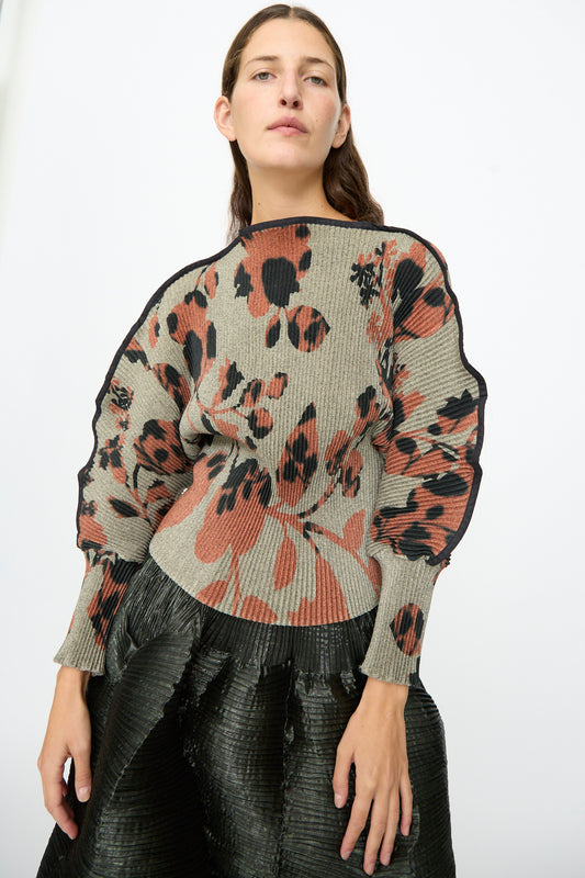 A woman wears the Julia Heuer Radok Pleated Knit Pullover in Ingrid—featuring abstract floral patterns in earthy tones and ribbed long sleeves—with a black textured skirt, standing against a plain white background.