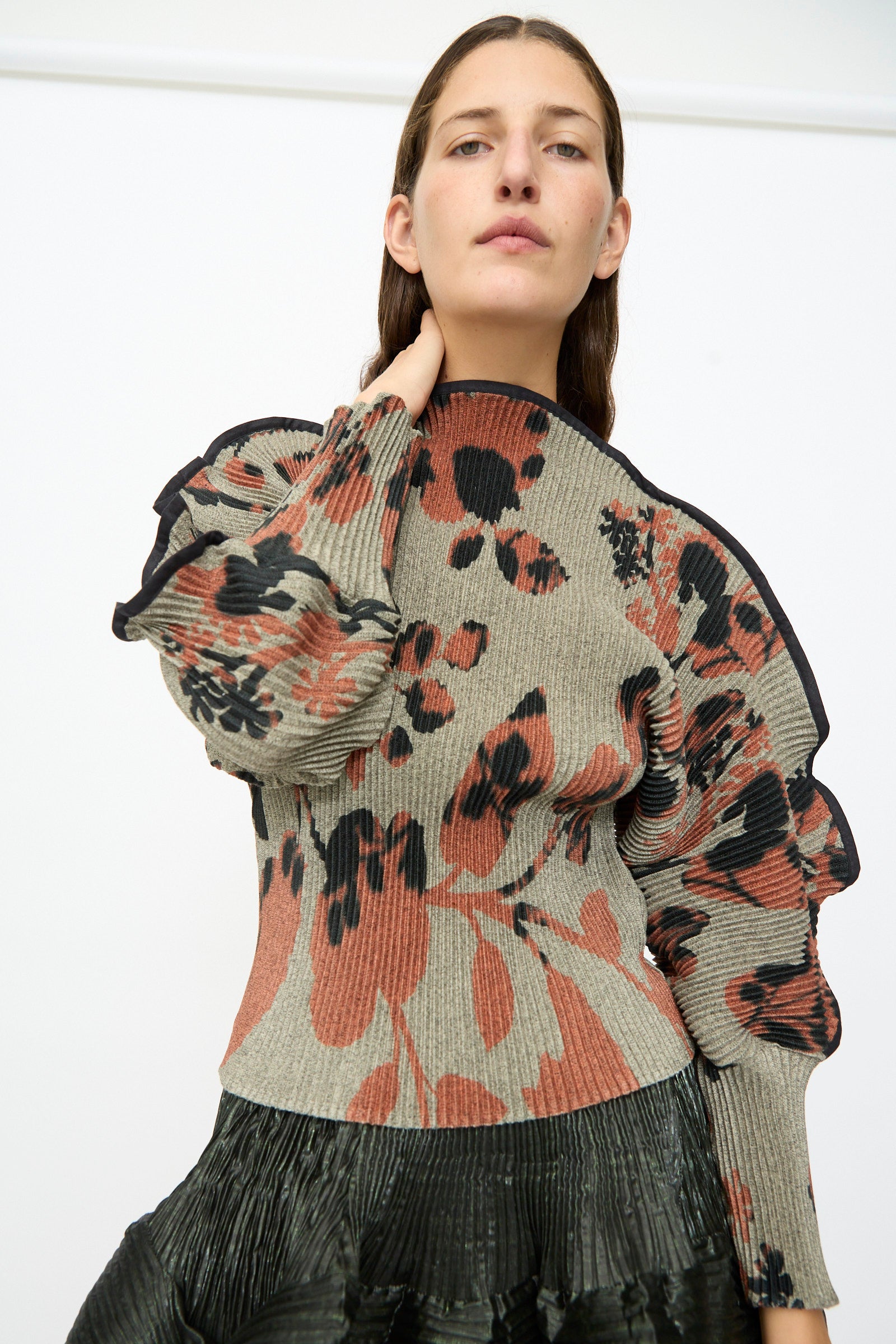 A person wearing the Julia Heuer Radok Pleated Knit Pullover in Ingrid, featuring gray pleats with black and rust floral patterns and sculpted long sleeves, paired with a pleated black skirt, poses against a plain white background.