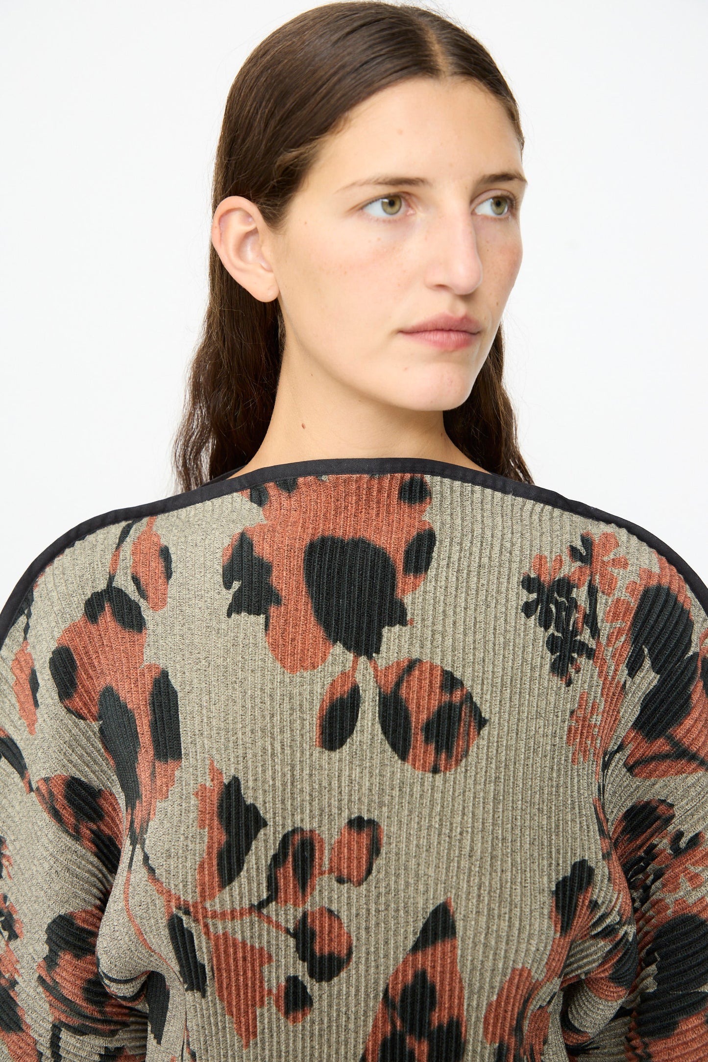 A woman with long brown hair wears the Julia Heuer Radok Pleated Knit Pullover in Ingrid, featuring a large abstract floral print in black and rust on a taupe background. This long-sleeve French-made sweater drapes elegantly as she gazes slightly to the right against a plain backdrop.