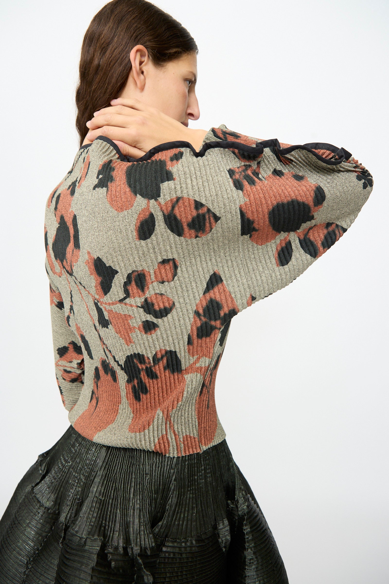 A woman faces away, wearing the Julia Heuer Radok Pleated Knit Pullover in Ingrid—an earth-toned, long sleeve top with a textured floral pattern and wide sleeves—paired with a high-waisted black pleated skirt. Made in France, she holds her neck with one hand.