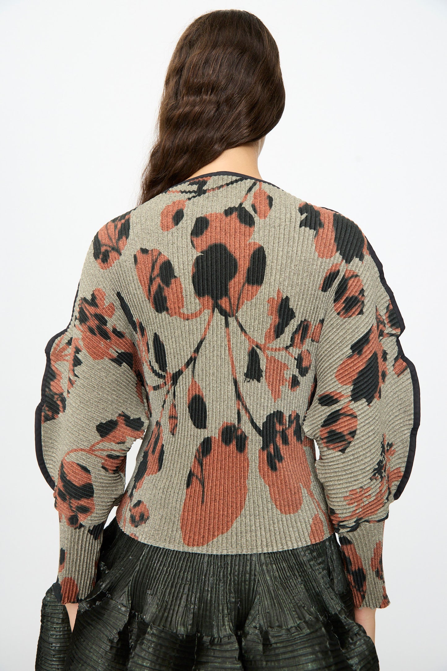 A person with long brown hair is shown from the back, wearing the Julia Heuer Radok Pleated Knit Pullover in Ingrid, featuring ribbed beige fabric with black and rust abstract patterns, paired with a pleated black skirt.