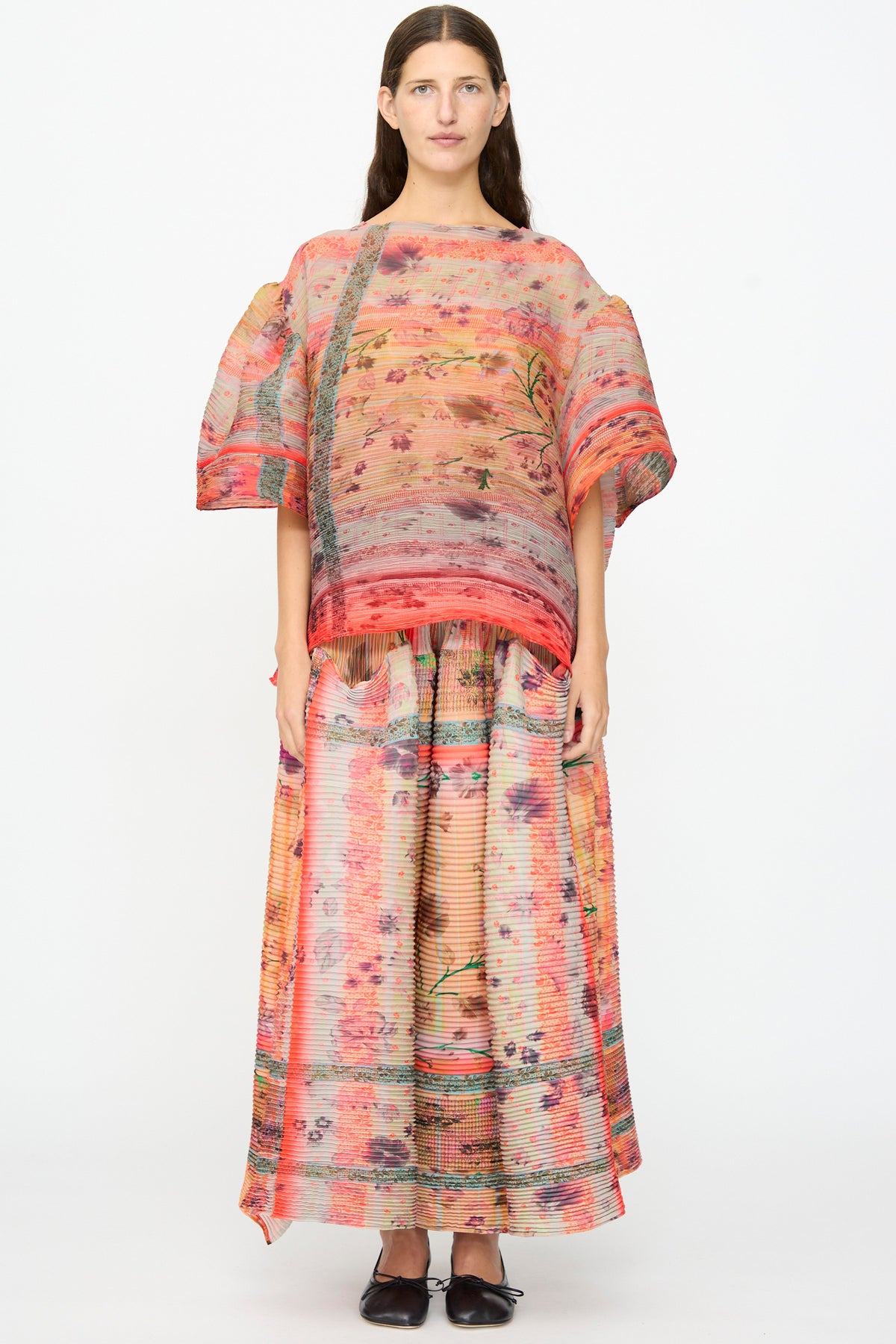 A woman stands against a plain background wearing the Julia Heuer Robin Handpleated Top in Bruno—an oversized, loose-patterned top with wide sleeves crafted from recycled chiffon. She pairs it with a matching long skirt, both featuring hand-pleated textures and abstract floral and striped designs in pink, orange, and beige tones.