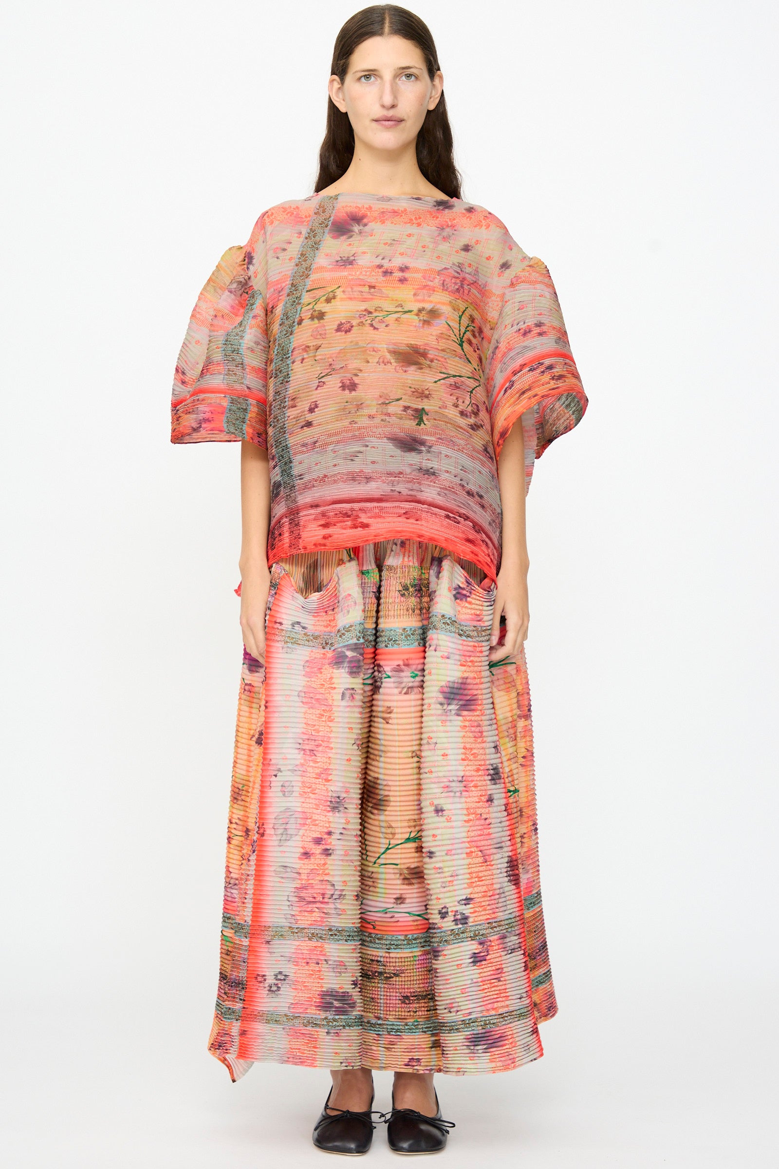 A woman stands against a plain background wearing the Julia Heuer Robin Handpleated Top in Bruno—an oversized, loose-patterned top with wide sleeves crafted from recycled chiffon. She pairs it with a matching long skirt, both featuring hand-pleated textures and abstract floral and striped designs in pink, orange, and beige tones.