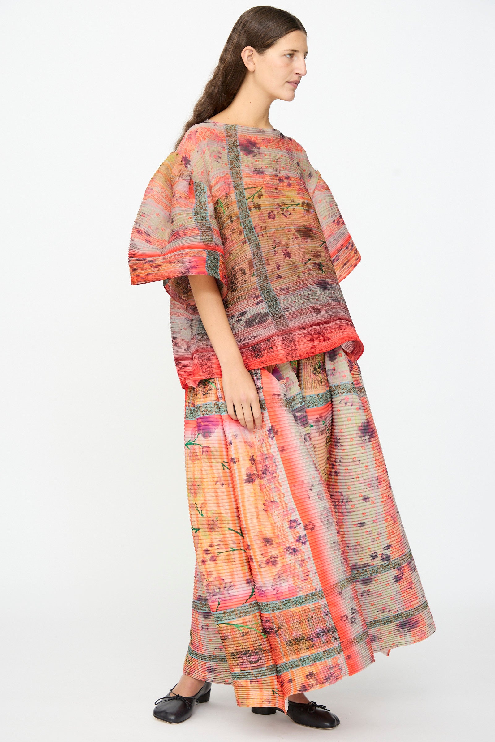 A woman stands sideways against a plain white background, wearing the Julia Heuer Robin Handpleated Top in Bruno—a loose, multicolored sculptural piece in an oversized fit—paired with a matching long skirt.