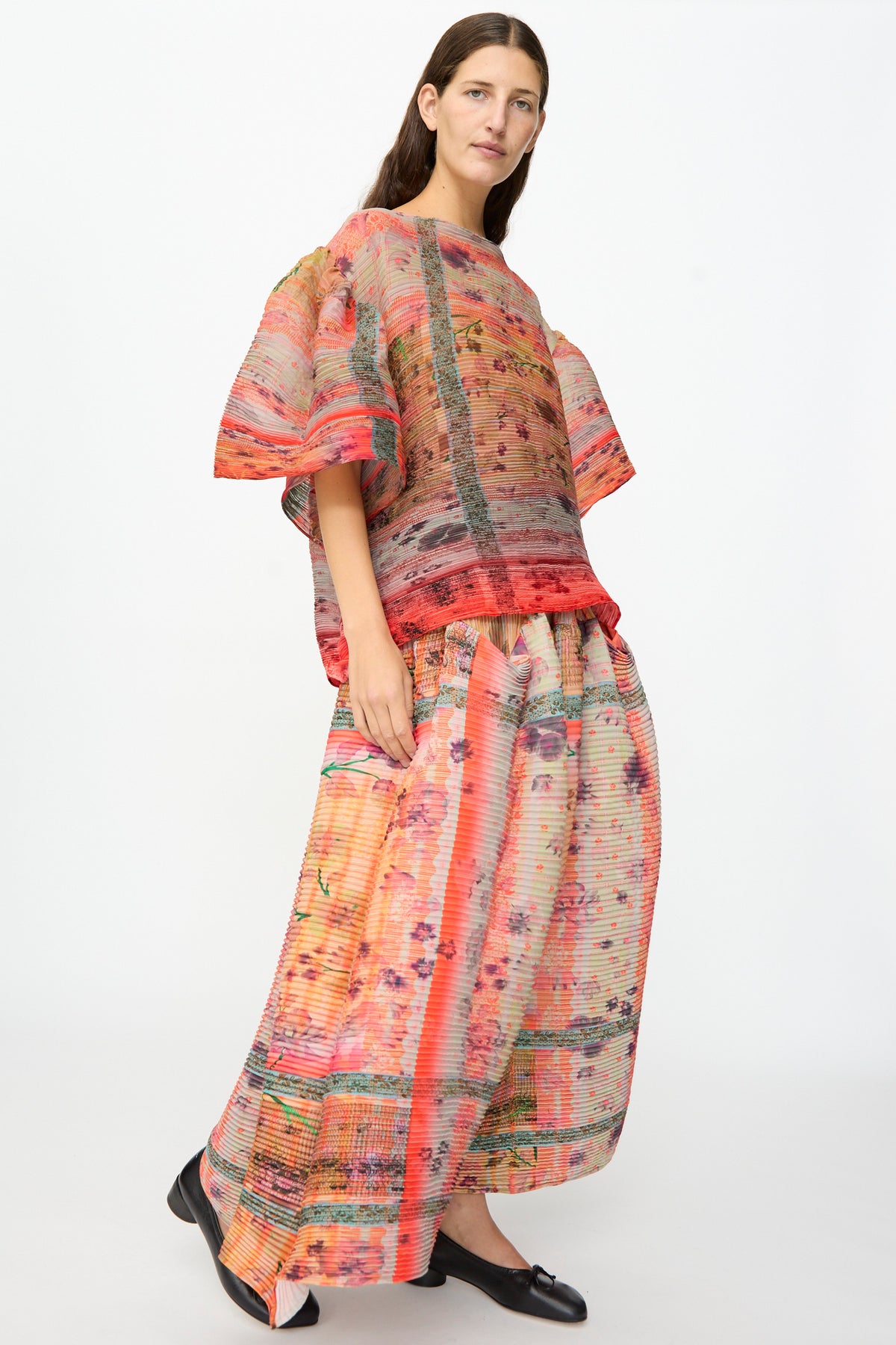 A woman wearing the Julia Heuer Robin Handpleated Top in Bruno—an oversized, colorful printed top with wide sleeves crafted from recycled chiffon—is paired with a matching long skirt and black shoes, standing against a plain white background.